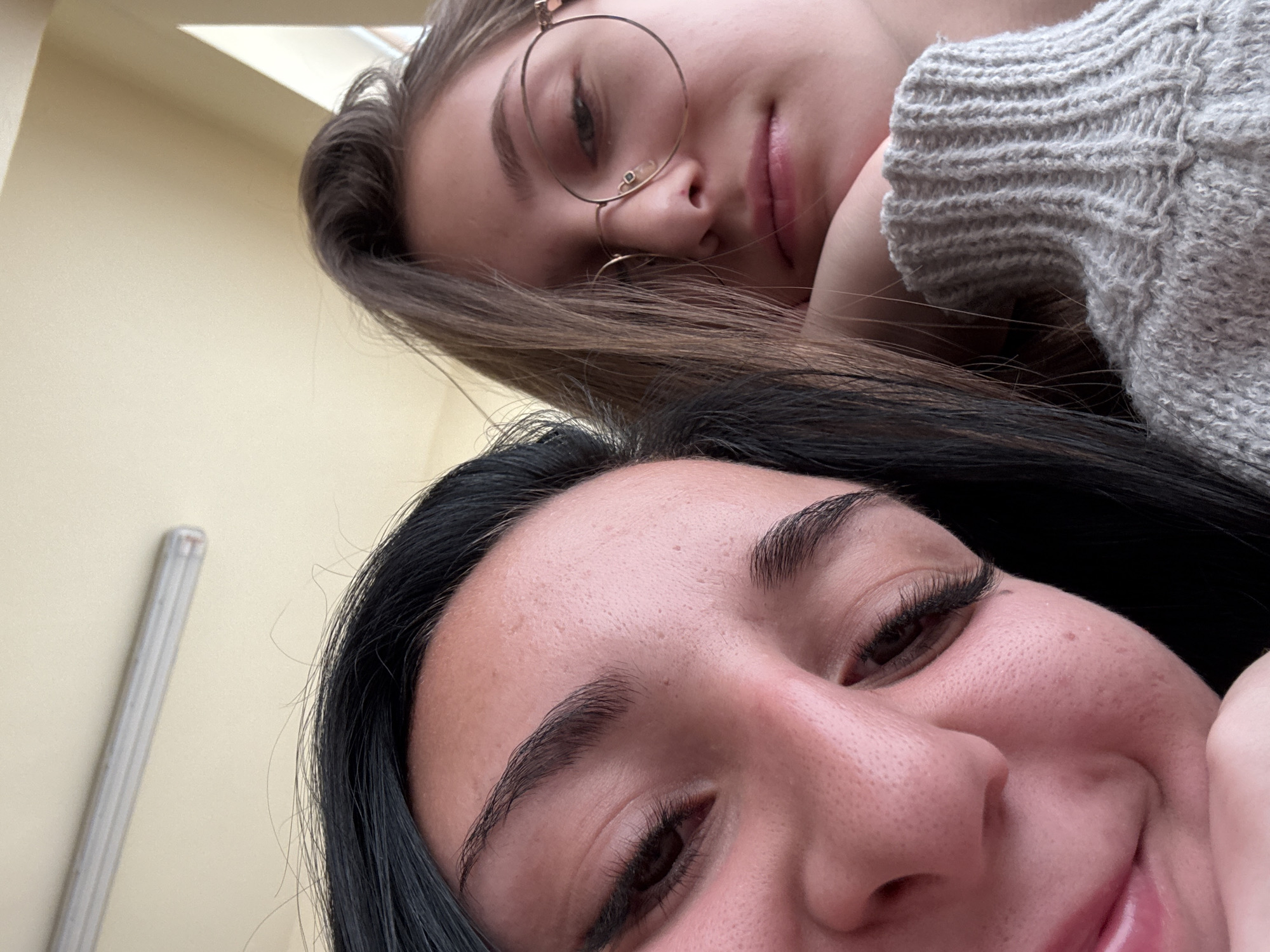 This close-up, angled photograph captures two young women indoors, likely posing for a selfie or self-shot. The image is taken from a low vantage point, looking upwards towards the subjects.
In the foreground, the face of a woman with dark, seemingly black hair is prominent. She has full, dark eyebrows and visible eyelashes, possibly extensions. Her skin shows some texture, and she is smiling warmly, with a hint of her pink lips visible. Her expression conveys cheerfulness.
Behind her, partially obscured, is another woman. She has lighter brown hair, which appears to flow towards the foreground. She wears distinctive round, wire-rimmed eyeglasses. Her expression is softer and more relaxed, with a gentle smile on her lips. She is dressed in what appears to be a light grey, textured, knitted garment, possibly a sweater.
The background consists primarily of a plain, light-colored wall or ceiling, possibly beige or off-white, suggesting an indoor setting. In the upper part of the background, there is an angular opening, possibly a skylight or a window, letting in natural daylight. Towards the lower left, a slim, silver-colored, vertically ridged object is visible against the wall, which could be part of a light fixture or vent.
The overall mood of the image is casual and friendly, capturing a moment between two individuals. The natural light suggests it is daytime. While the provided context states Sofia, Bulgaria, there are no specific visual cues within the image itself to identify this location. No text is visible in the photograph.