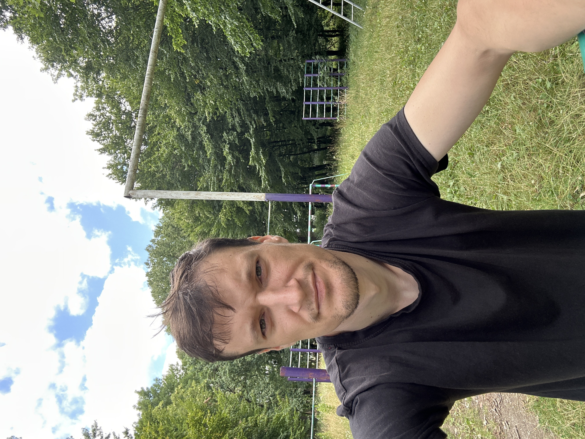 A mid-shot, selfie-style photograph captures a man with short, dark brown hair and a light beard, wearing a black t-shirt. He has a neutral expression and is looking directly at the camera. The background is an outdoor setting with a grassy area and a dense cluster of green trees. In the mid-ground, there are elements of an outdoor gym or playground, including purple and white posts, horizontal bars, and a ladder. A portion of a white horizontal bar, likely part of a goalpost or similar structure, is visible in the upper right quadrant. The sky above is partially obscured by clouds. The scene is set in the city of Makiv, Ukraine.