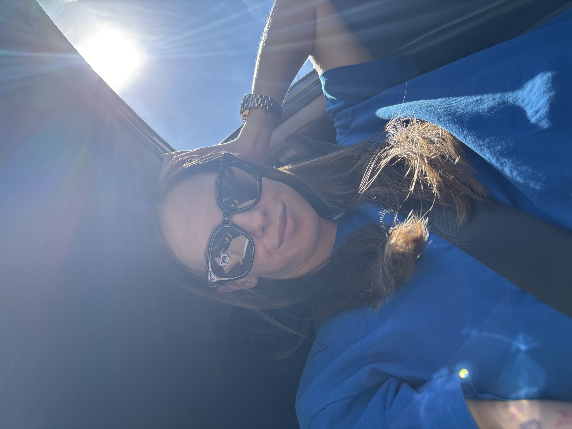 The media file is a selfie taken by a young woman, who is the main subject. She is wearing a bright blue t-shirt and sunglasses, and her brown hair is long and partially covering her face. A silver watch is visible on her wrist. The setting appears to be inside a vehicle, possibly with a sunroof open, as evidenced by the bright sunlight streaming in and the angle of the shot. The strong sunlight suggests it is daytime, likely mid-morning or afternoon. The woman is looking towards the camera, with a neutral or slightly smiling expression. The background is mostly out of focus, but hints of the sky and interior of a car are discernible. The overall mood is casual and bright. There is some text visible on the woman's t-shirt, but it is largely obscured by hair and lighting conditions, appearing to be a small logo or inscription.