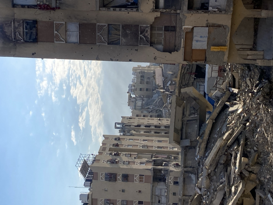 The image captures a scene of extensive urban devastation under a clear blue sky with scattered white clouds. The perspective appears to be from a vantage point within or near a damaged structure, looking out over a ruined cityscape.
In the immediate foreground, likely above the viewer, is a severely damaged concrete structure. It has multiple rectangular openings, some of which appear to be windows that are broken, boarded up with various materials (some brown, some white fabric-like), or simply gaping holes revealing darkness or debris inside. The concrete surface shows numerous small impact marks or holes, suggesting shrapnel damage or bullet holes. On a section of exposed wall or debris within this damaged structure, a partially visible "UNICEF" logo in blue and white is discernible.
Below and beyond this foreground structure, a wide expanse of destroyed and damaged buildings is visible. On the left side of the frame, several multi-story residential or commercial buildings stand. Many of these structures exhibit significant damage, including broken windows, missing sections of walls or balconies, and some blackened areas that might indicate fire damage. One building on the far left has a large, empty billboard-like frame on its roof.
The right side and central midground of the image are dominated by a massive pile of rubble. This debris consists of large concrete slabs, broken bricks, twisted metal, and other construction materials, indicating the complete collapse of one or more buildings. Behind and amidst this rubble, other buildings are seen in various states of disrepair and partial collapse. One building appears to have had its upper floors entirely destroyed, leaving only lower sections and a chaotic jumble of internal structures.
The overall scene conveys the aftermath of a catastrophic event, such as a major conflict or a natural disaster like an earthquake. The bright daylight, with clear blue skies and wispy white clouds, provides a stark contrast to the widespread destruction below. There are no people visible, and no active events are taking place; the image is a static portrayal of devastation.