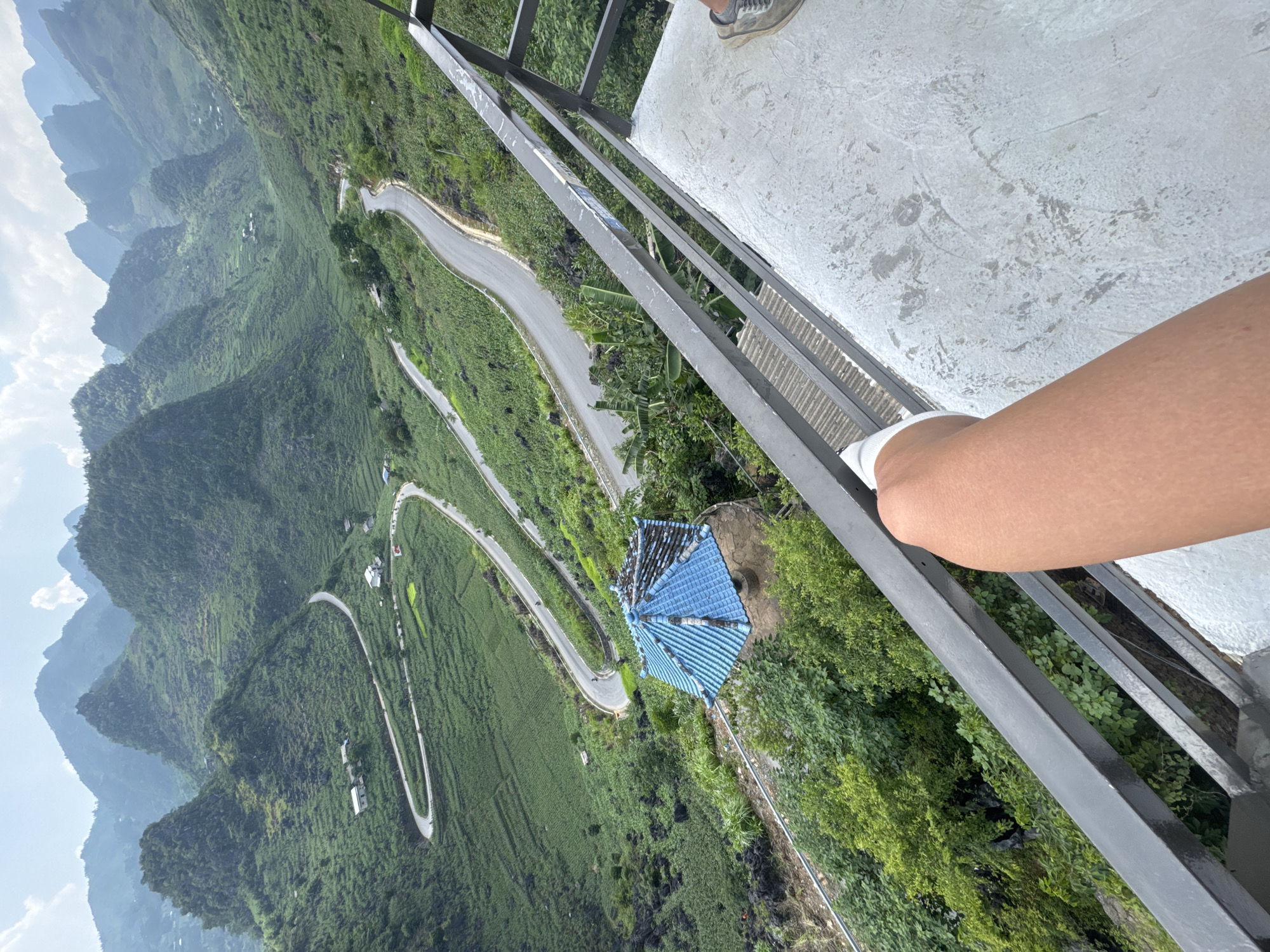 An elevated shot overlooks a sinuous mountain road near Lũng Phìn Commune, Vietnam. The shot is taken from a high viewpoint, likely a structure with a gray metal railing and a concrete base, visible in the frame. A leg wearing a white sock rests against the railing. Below, the landscape is characterized by verdant hills, terraced fields, and scattered buildings. A structure with a prominent blue tiled roof stands out. The road winds through the landscape, ascending and descending the hills. In the distance, layers of mountains are visible under a sky with scattered clouds.