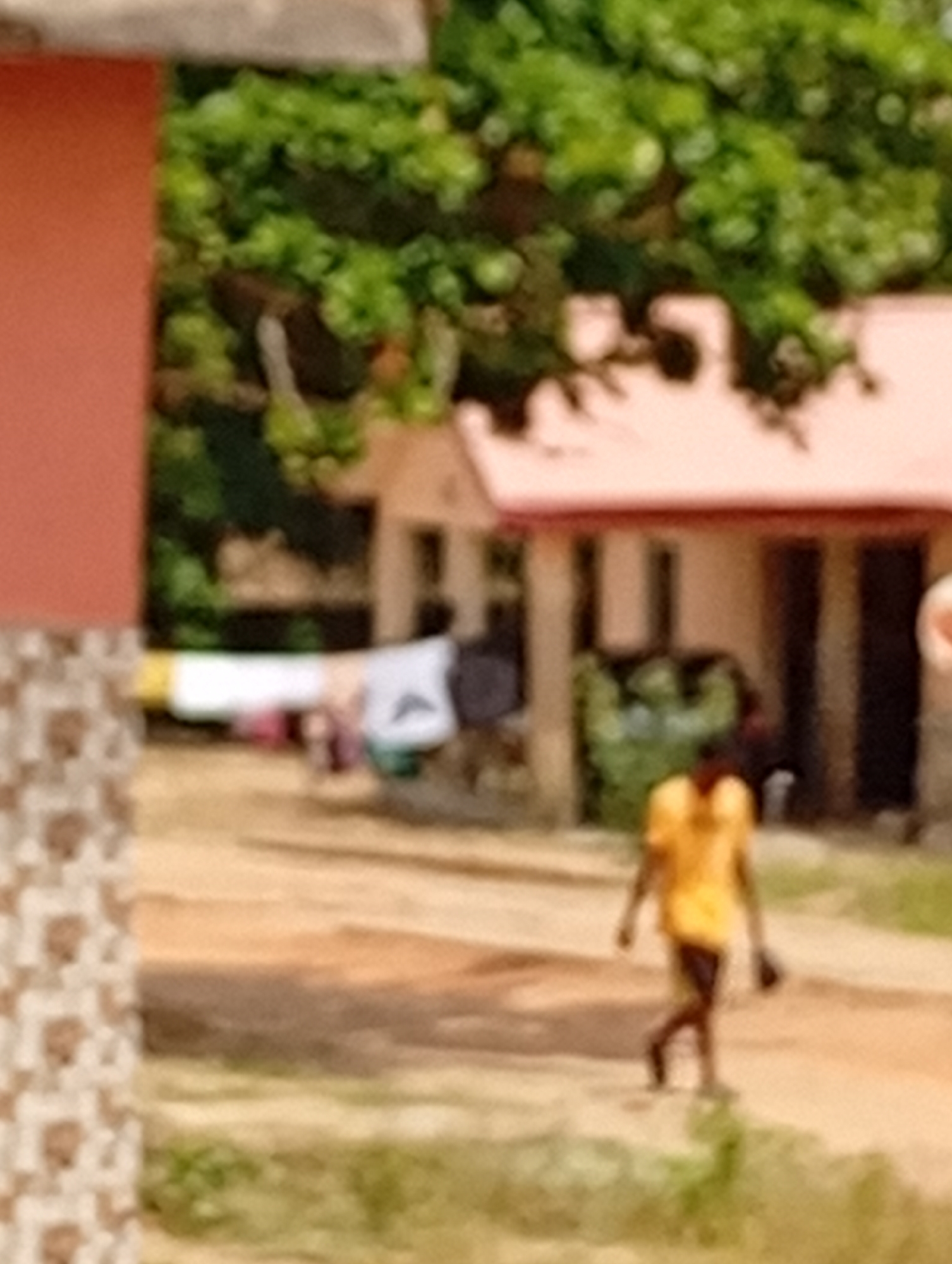A blurred scene captured in an unspecified location in the city of Unknown, Unknown shows a person in a yellow shirt walking on a dirt road. In the background stands a house with a red roof, partially obscured by trees. Clothing is hanging on a line between the house and the camera. Buildings are seen on either side of the scene.