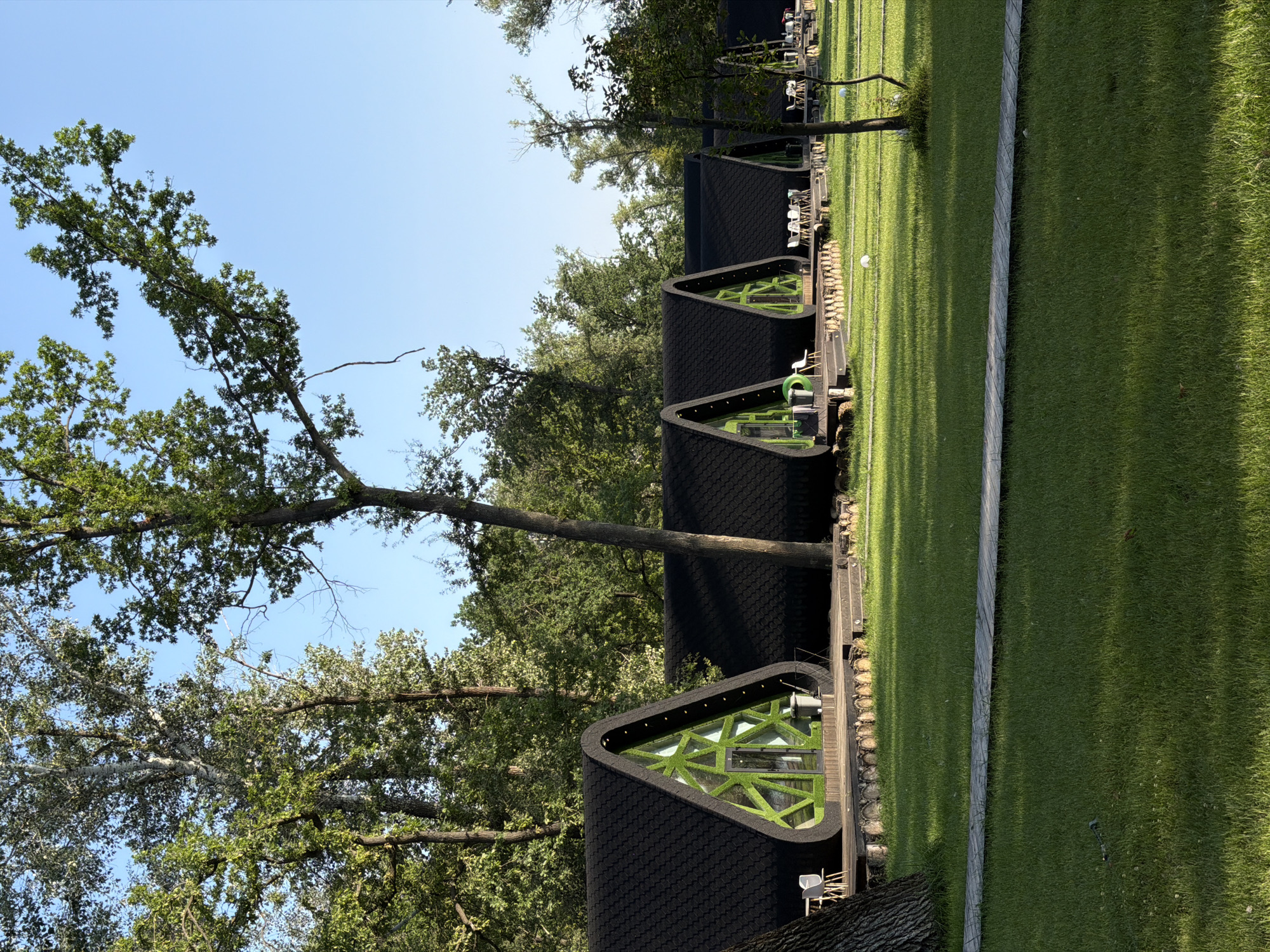 The image depicts a row of modern, triangular cabins with black, textured roofs and distinctive green, geometric window facades. These cabins are situated on wooden decks, with a manicured lawn in front of them. Several white chairs and tables are visible on the decks, suggesting an outdoor seating area. A wooden walkway runs across the lawn. In the background, lush green trees and a clear blue sky form the natural environment. The scene appears to be a resort or a glamping site. The location is identified as being in the city of Engels, Russia.