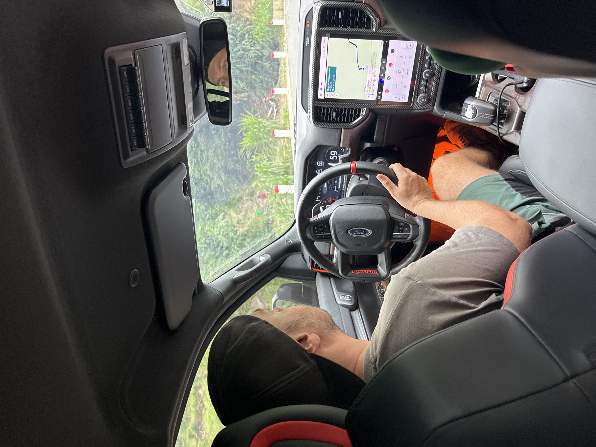The image captures the interior of a modern Ford Raptor vehicle, viewed from a rear passenger seat looking forward. The photo is rotated 90 degrees clockwise. A male driver, wearing a dark cap, grey t-shirt, green shorts, and orange shoes, is seated in the driver's seat, holding the steering wheel with his right hand.
The dashboard features a prominent, vertically oriented central infotainment screen displaying Google Maps navigation, showing a route with an estimated time of 13:02 and a temperature of 22.5°C. Climate controls are visible below the screen. The digital instrument cluster behind the steering wheel shows "59," likely a speed reading. The steering wheel is branded with "FORD" and "RAPTOR."
An overhead console contains six auxiliary switches labeled "AUX1" through "AUX6." The rearview mirror reflects a person, possibly female, wearing a dark head covering, suggesting the photographer. Through the windshield, green foliage and several red and white striped poles are visible, indicating an outdoor setting, potentially a path or trail in Minh Sơn Commune, Vietnam. The gear shifter is a rotary dial. The vehicle's interior is dark-toned with black seats featuring red accents. The scene takes place during bright daylight.