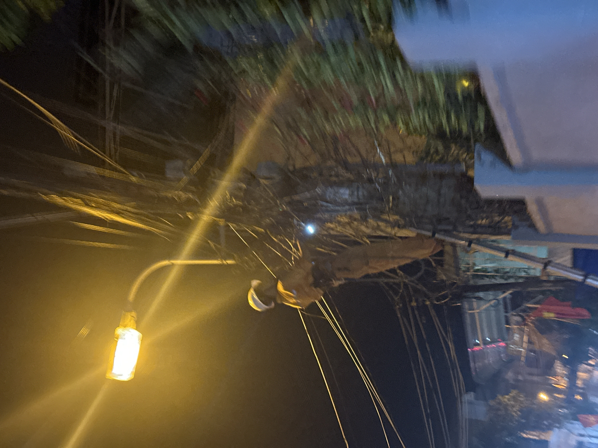 Captured at night, the photograph shows an outdoor scene dominated by a dense network of utility wires converging near a street light, illuminating the area with a yellow glow. A worker wearing a helmet appears to be attending to the wires. In the background, architectural elements and structures can be discerned, along with the faint outline of a flag, suggesting a Vietnamese context. It is located in Bac Nha Trang Ward, Vietnam.