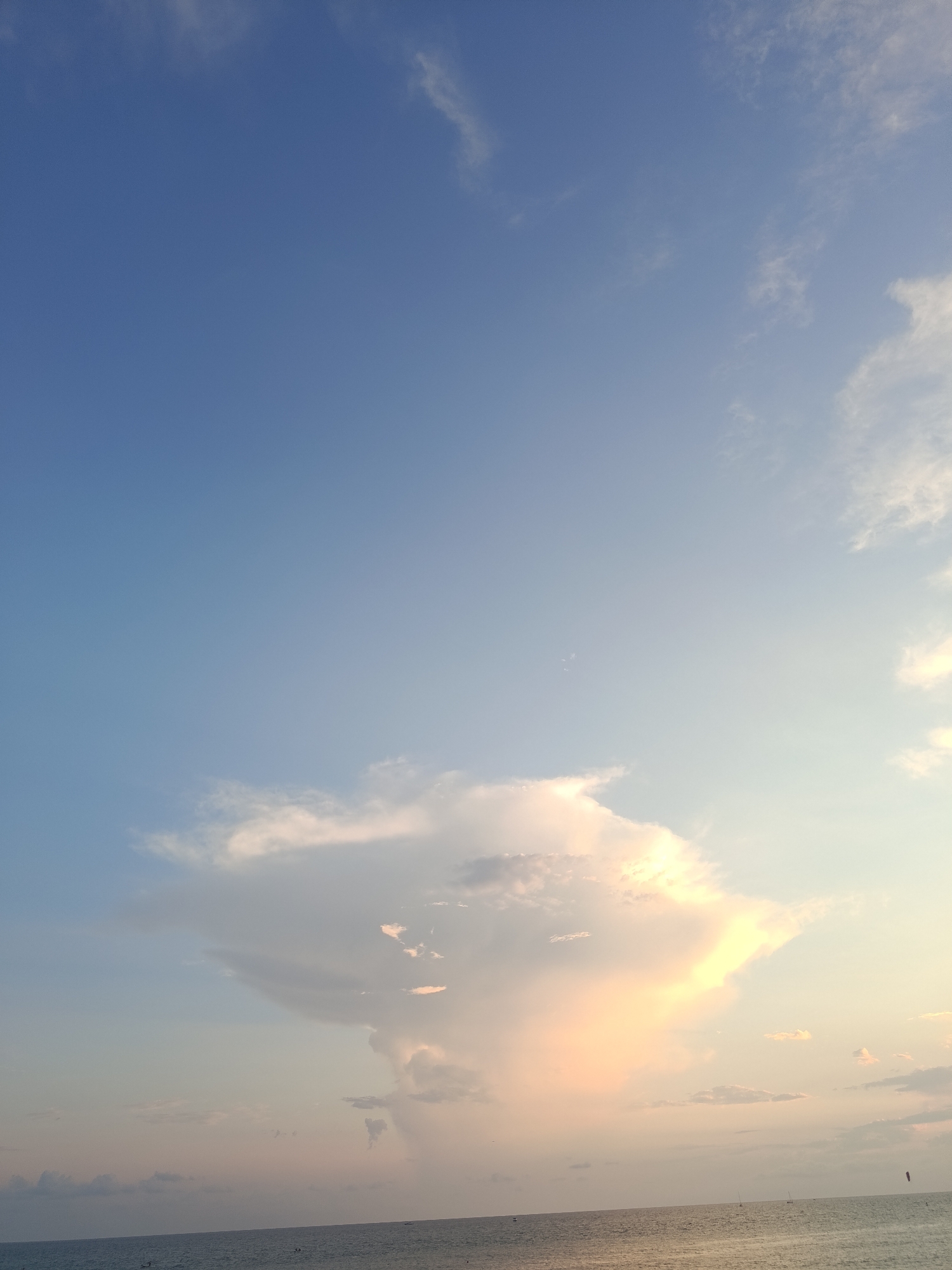 A wide shot captures the tranquil expanse of the Black Sea, with a calm, dark blue body of water stretching across the bottom of the frame. A thin, lighter-colored horizon line separates the water from the sky. The sky above is a gradient of soft blues, transitioning to a pale, warm yellow and pinkish hue near the horizon, suggesting the presence of a setting sun. A large, dramatic cloud formation dominates the center of the sky, illuminated by the warm light, creating a striking contrast against the bluer parts of the sky. The cloud has intricate textures and appears to be catching the light from below. Faint, wispy clouds are scattered throughout the upper portion of the sky. A few small, dark shapes are visible on the water's surface, possibly distant boats or buoys. The scene is serene and evokes a sense of natural beauty and peacefulness. The location is identified as Sirius, Russia.