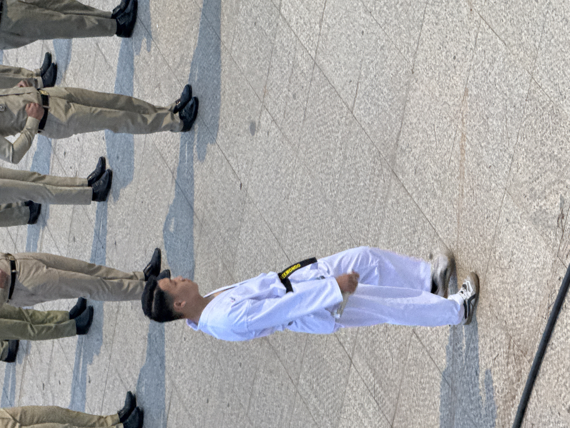 The image depicts a group of individuals, likely students or cadets, standing in formation on a paved outdoor area. The majority are wearing light-colored uniforms with dark shoes. In the foreground, a young man wearing a white karate gi with a black belt that has yellow lettering, possibly "TAEKWONDO," stands facing to the left. He appears to be holding a white object in his right hand. The scene is likely set in Da Lat, Vietnam, given the tiled ground that is characteristic of many public spaces and walkways in the city. The lighting suggests it is daytime, with shadows cast on the ground, indicating sunlight.