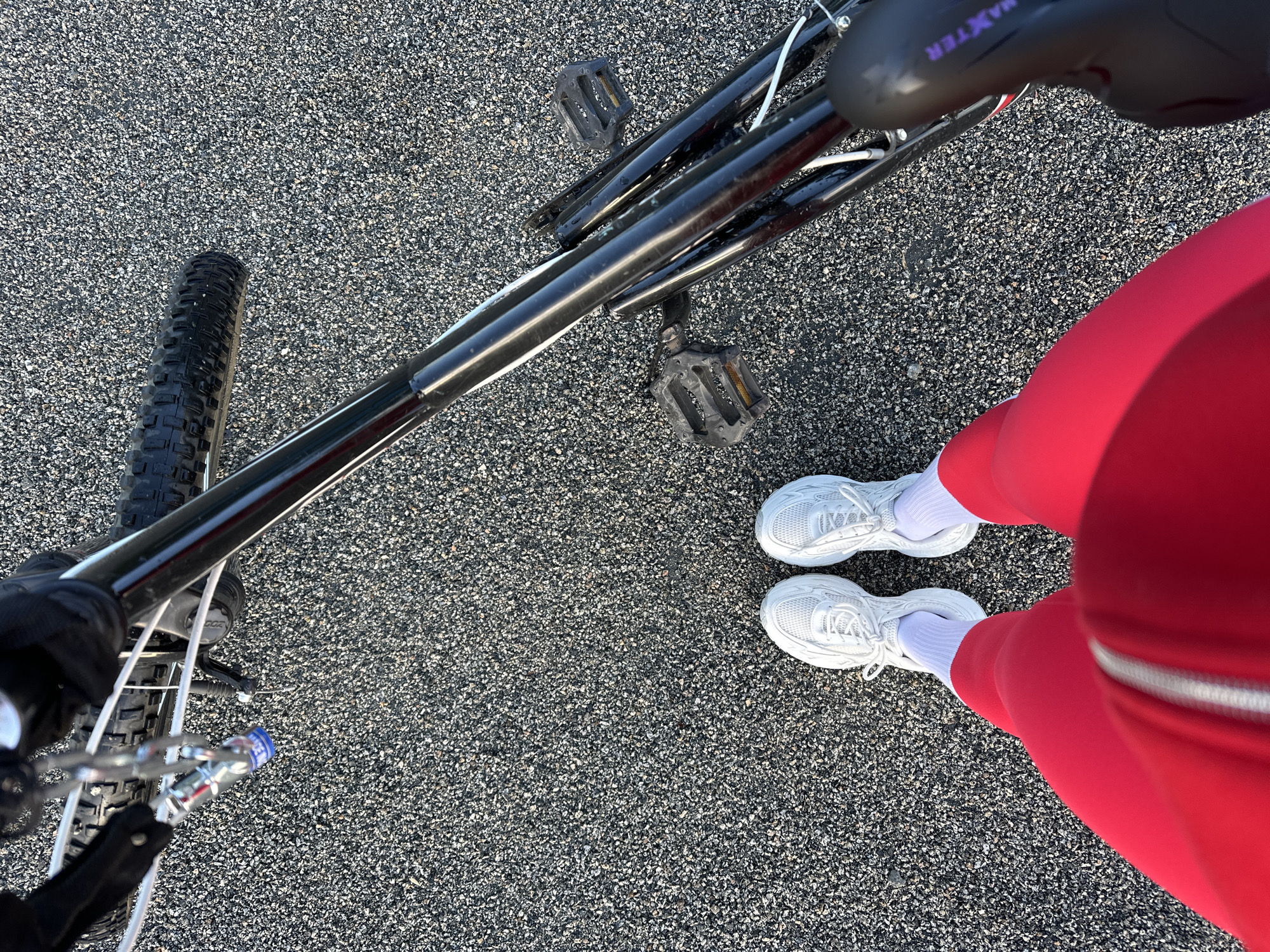 The image, taken from a high-angle, first-person perspective, depicts a person standing next to a bicycle on a dark, granular asphalt surface. The individual wears red athletic pants with a visible white zipper, white ankle socks, and white chunky-soled athletic sneakers. A black-framed bicycle is positioned to the person's right. It features knobby tires, black platform pedals, and a black saddle inscribed with "MAXTER" in purple lettering. A silver chain lock, with a blue cap partially reading "LICE", hangs from the bicycle's front section. White brake cables run along the frame, which displays minor wear. The ground is dark grey asphalt, densely paved with small, lighter-colored gravel. The scene is brightly lit by direct sunlight, creating clear shadows. This scene is located in Yuzhnouralsk, Russia.