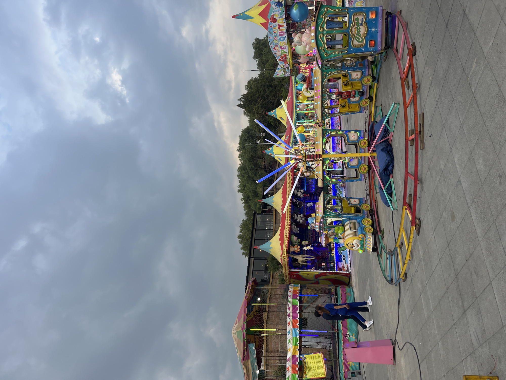 The image captures a children's amusement ride set up outdoors in what appears to be a temporary fair or carnival setting, presumably located in San Andrés Cholula, Mexico. The ride features a circular track with brightly colored, cartoon-themed carriages, and illuminated with festive lights. Various tent structures and colorful signage indicative of a fair are visible in the background. In the foreground, two individuals stand beside a brightly colored barrier. The sky is overcast with heavy clouds. The ground is paved with gray tiles.