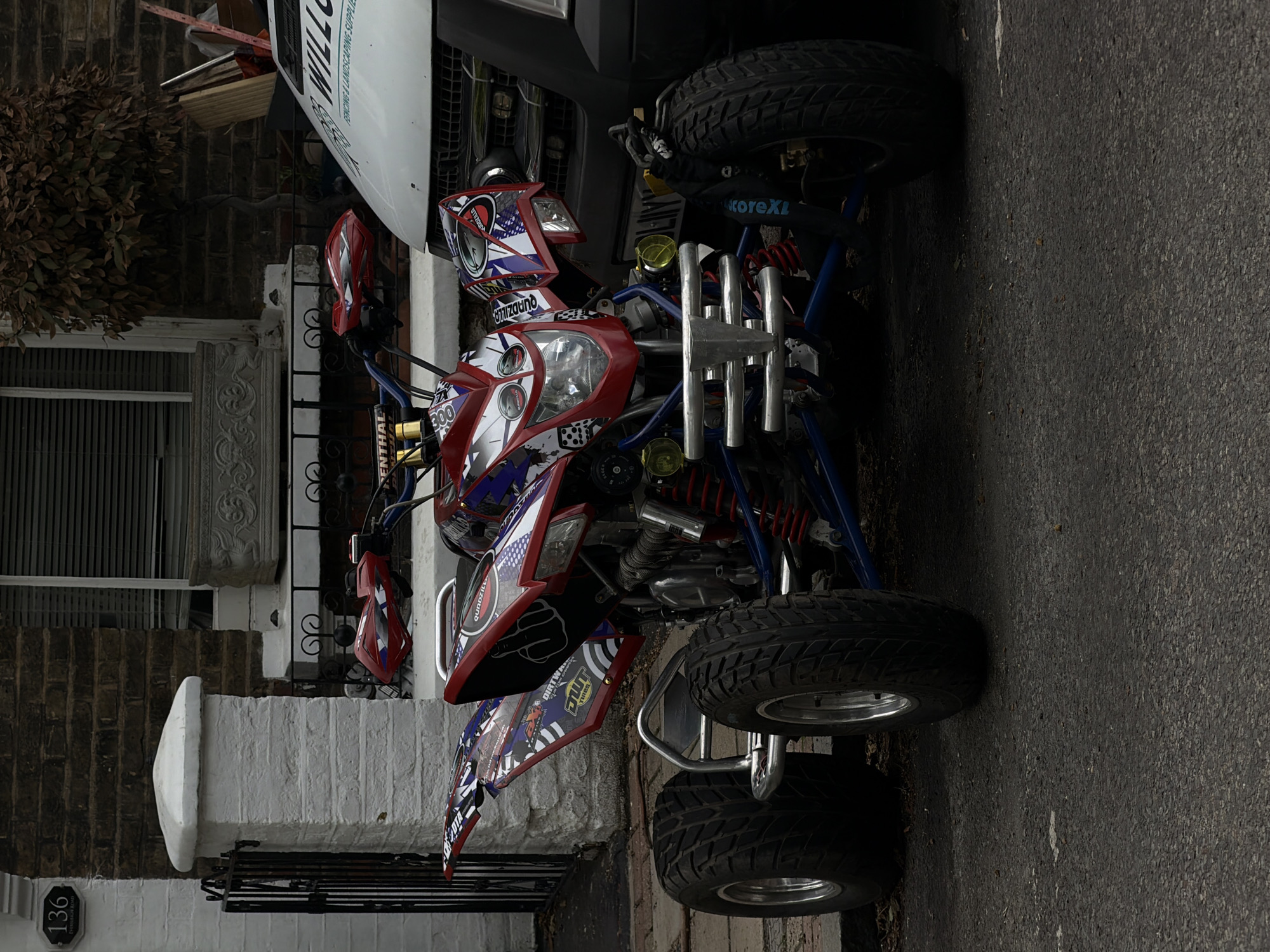 A red, white, and blue patterned quad bike is parked on an asphalt surface in front of a brick building with white trim. The quad bike has yellow headlights and a chrome bumper. A white van with "WILLOW" and "FENCING & LANDSCAPING SUPPLIES" written on it is parked to the right of the quad bike. The building has a black metal fence and a white pillar with a house number "136" on it. There is a tree with brown leaves to the right of the building. The scene appears to be in a residential area in London, United Kingdom.