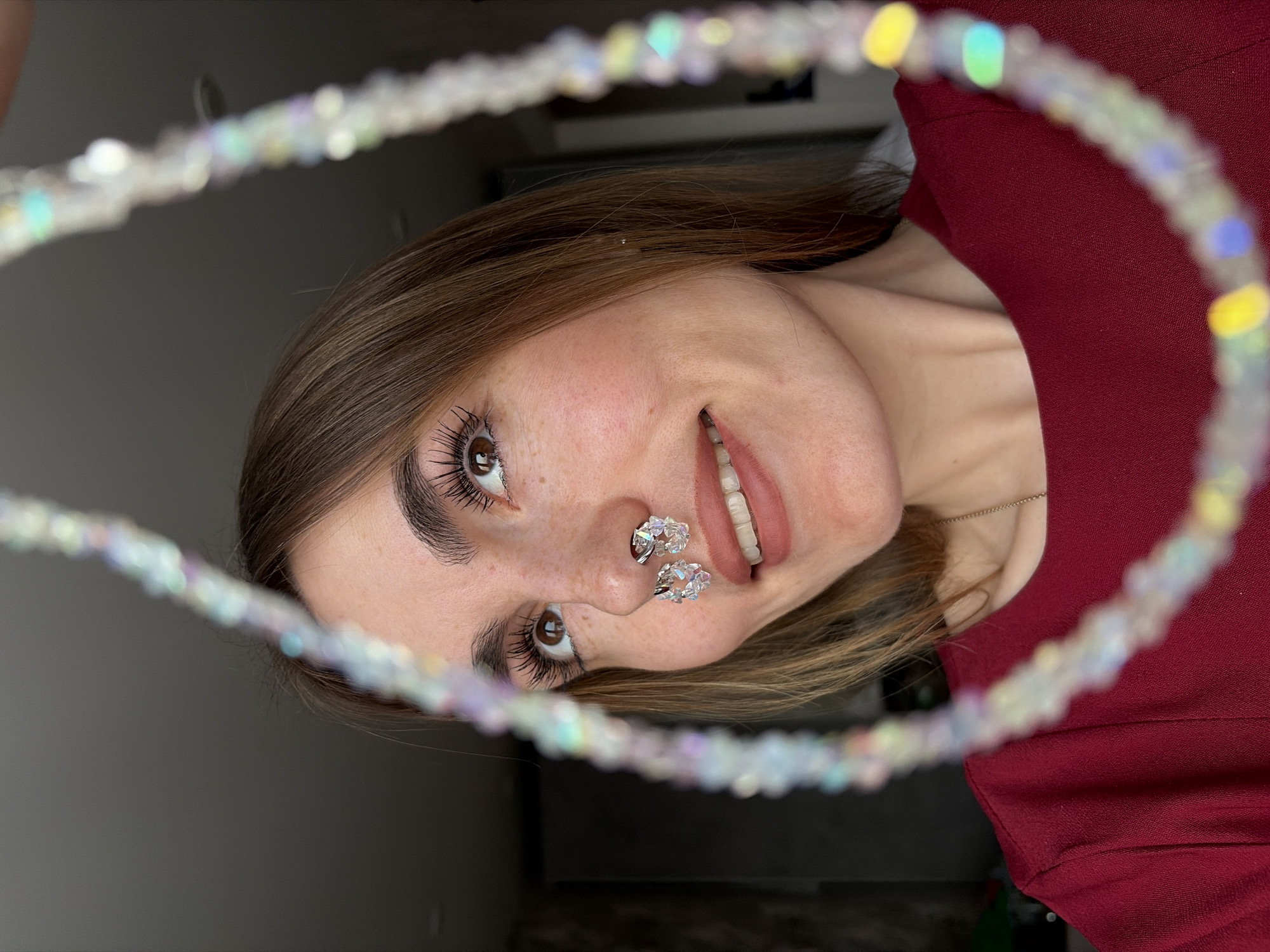 A woman with brown hair and eyes, fair skin, and freckles smiles while holding a blurry, beaded halo in front of her face. She is wearing a burgundy shirt. She has two large, crystal-like nose rings. The background is a light gray wall. This image does not provide sufficient information to determine if it was taken in Izhevsk, Russia.