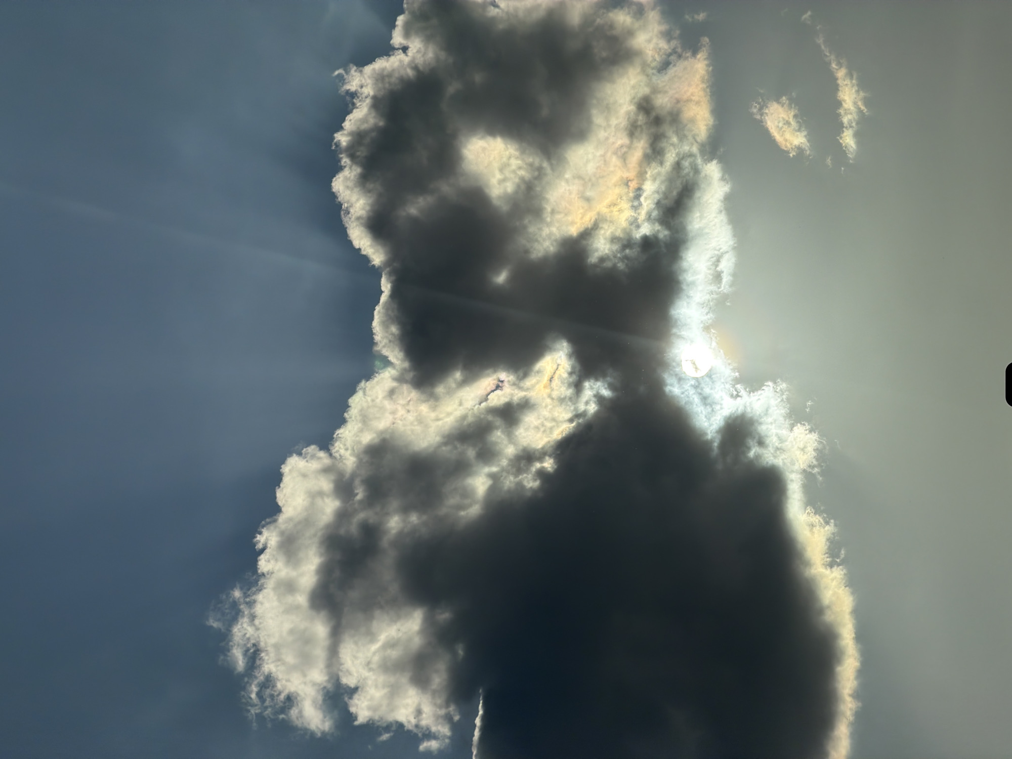 The image is an outdoor shot of a cloudy sky. The sun is partially obscured by a large, prominent cloud, casting rays of light around it. The cloud has dark and light areas, creating a dynamic contrast. The sky is blue on the left side of the image and becomes lighter near the sun. There are a few smaller clouds scattered in the sky. The image is taken at an angle, with the clouds appearing vertically. The image was captured in Esch-sur-Alzette, Luxembourg.