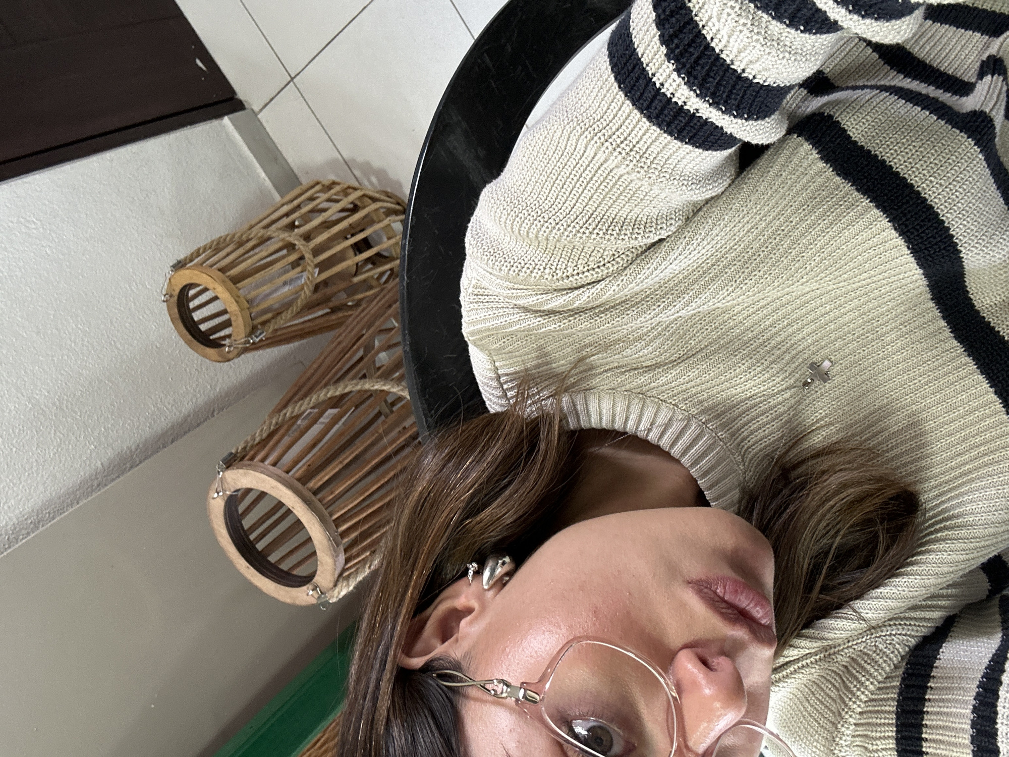 A close-up, rotated view shows a woman wearing glasses and a striped sweater, sitting at a black table. Two wooden lanterns are positioned on a surface near the table. The location is Unknown, Unknown.