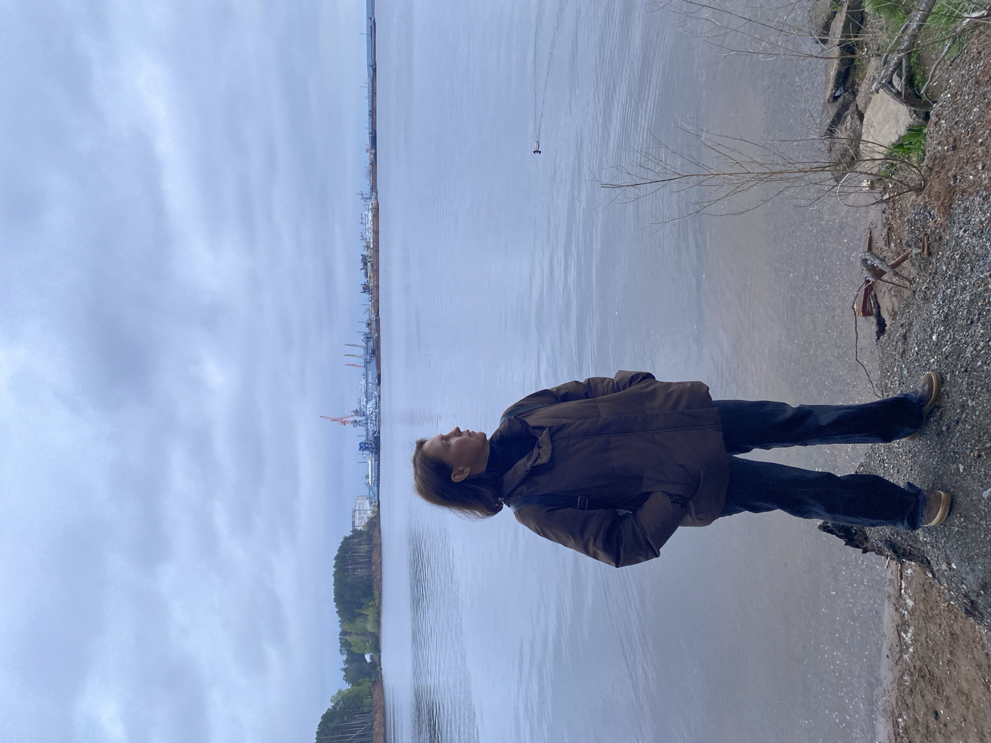 A person stands on the shore of the Kama River in the city of Chaikovsky, Russia. The person is wearing a brown jacket, dark jeans, and brown shoes. They are looking out at the river. In the background, there are ships and cranes at the Chaikovsky shipyard. The sky is overcast and the water is gray. There is a small bird swimming in the river.
