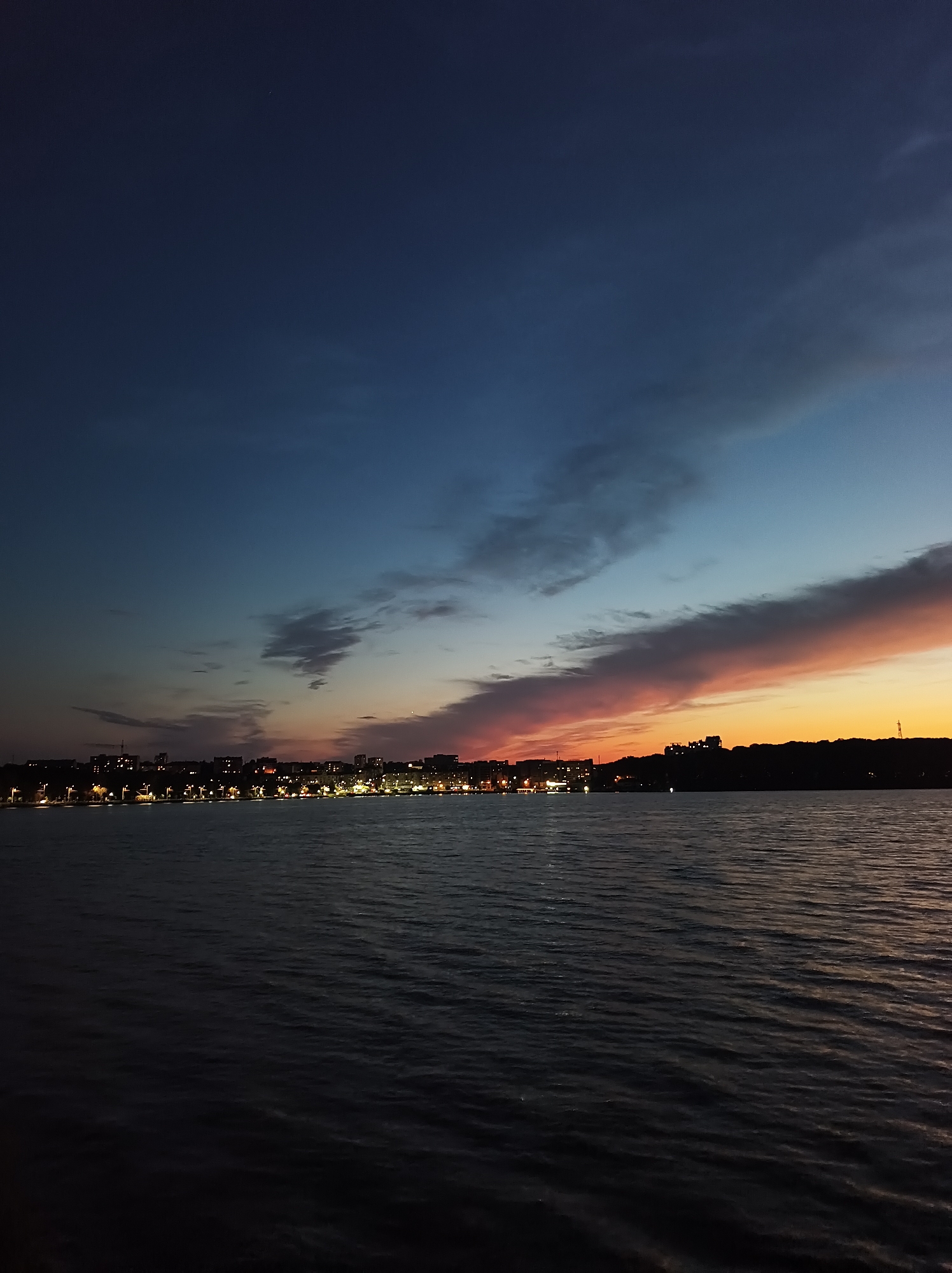 The image depicts a panoramic view of the Ternopil Reservoir in Ternopil, Ukraine, during twilight. The sky is a gradient of deep blue transitioning to hues of orange and pink, with scattered clouds catching the last rays of the setting sun. A line of city lights illuminates the distant shoreline, suggesting a populated area. Buildings of varying heights are visible, silhouetted against the fading light. The foreground is dominated by the dark, rippling surface of the water, reflecting the muted colors of the sky. The overall scene is tranquil and atmospheric, capturing the serene beauty of the reservoir at dusk. There are no people or specific objects of interest in the foreground or mid-ground, focusing solely on the natural and urban landscape.
