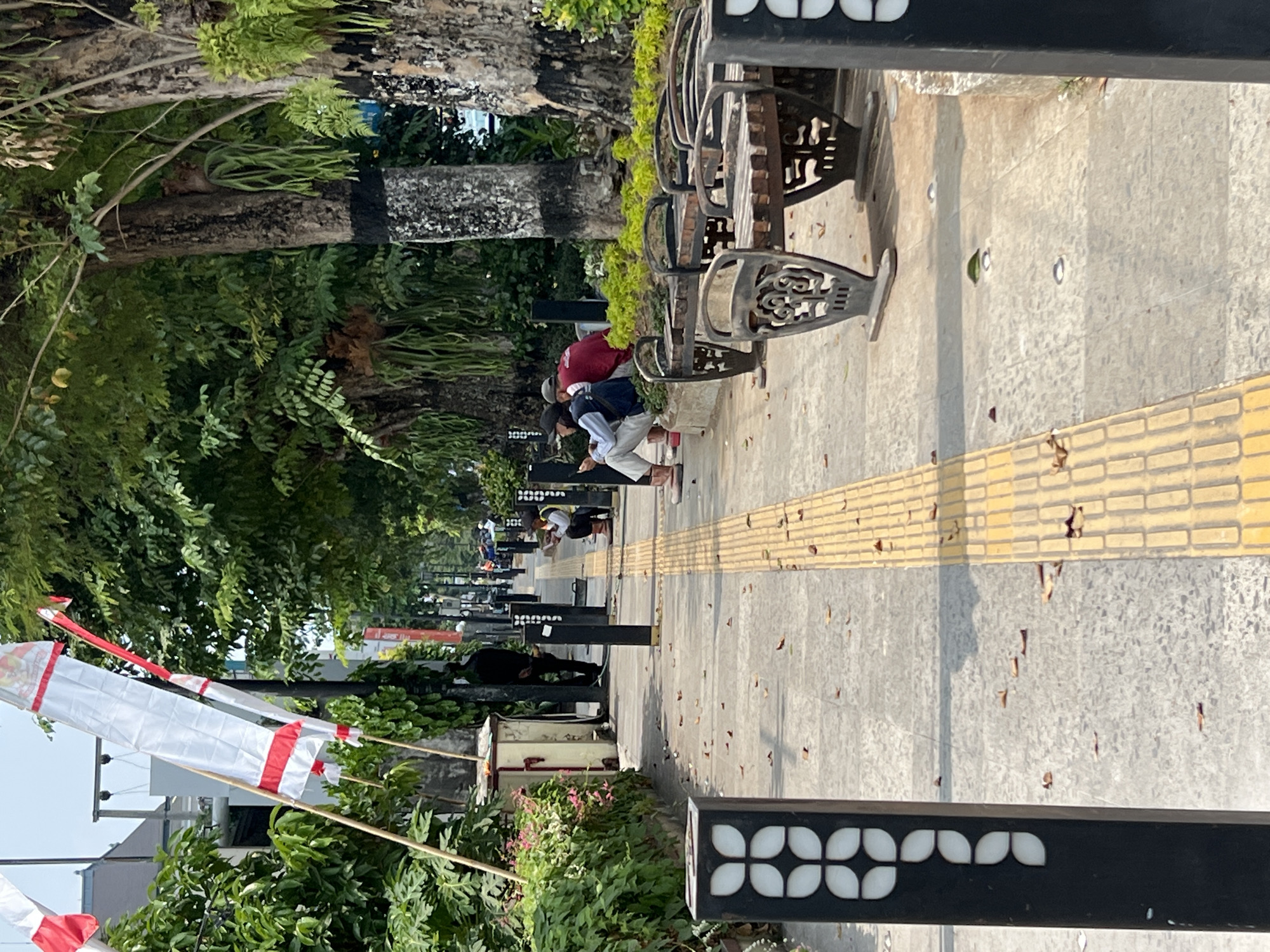 The image depicts a pedestrian sidewalk in a city, likely in Yogyakarta, Indonesia, judging by the red and white flags adorning poles, which are reminiscent of the Indonesian national flag. The sidewalk is paved with concrete and features a textured yellow strip, possibly a tactile paving system for the visually impaired, running along its length. Several people are present on the sidewalk. In the foreground, a black bollard with a white, four-leaf clover-like design is visible on the left. Further down the sidewalk, two individuals are seated on a park bench, with one looking at their phone. Behind them, a person in a dark jacket stands near a trash receptacle. More people are visible further in the distance, suggesting a public space or thoroughfare. Lush green trees with thick trunks line the right side of the sidewalk, casting shadows. Some of the trees have ferns growing on them. The scene is bathed in daylight, indicating it is daytime. The atmosphere appears relaxed, with individuals engaged in their own activities.