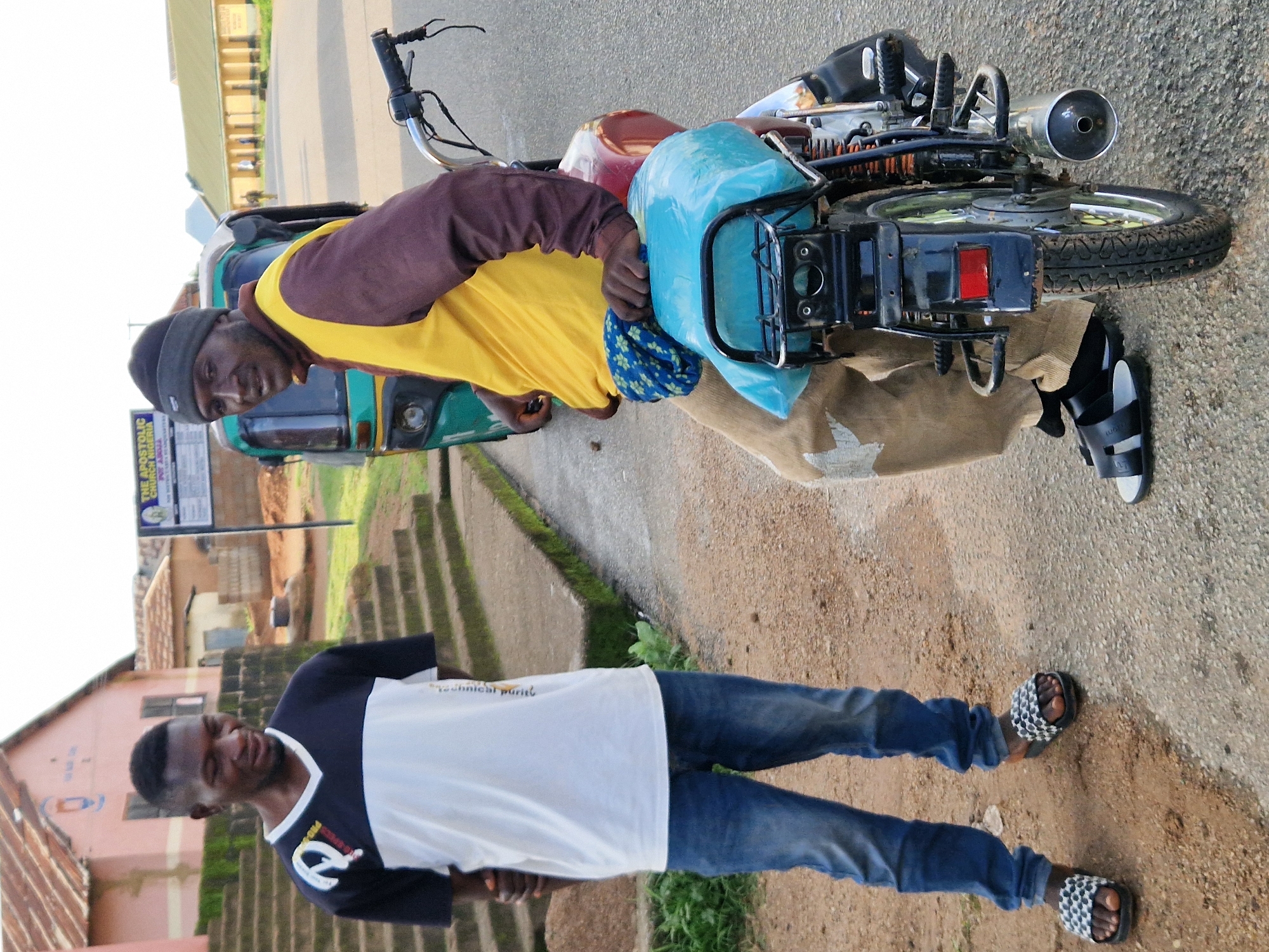 Two men are standing next to a motorized tricycle and a motorcycle on a paved road in Kabong, Nigeria. The man on the left is wearing a white and black t-shirt, blue jeans, and patterned slide sandals. He has his hands clasped behind his back. The man on the right is wearing a dark beanie, a maroon long-sleeved shirt, a yellow vest, and light brown pants. He is seated on the motorcycle. The motorcycle has a blue tarp covering its seat and a chrome exhaust pipe. In the background, there are buildings, a sign that reads "THE APOSTOLIC CHURCH NIGERIA," and a set of concrete stairs. The environment appears to be dry and dusty.