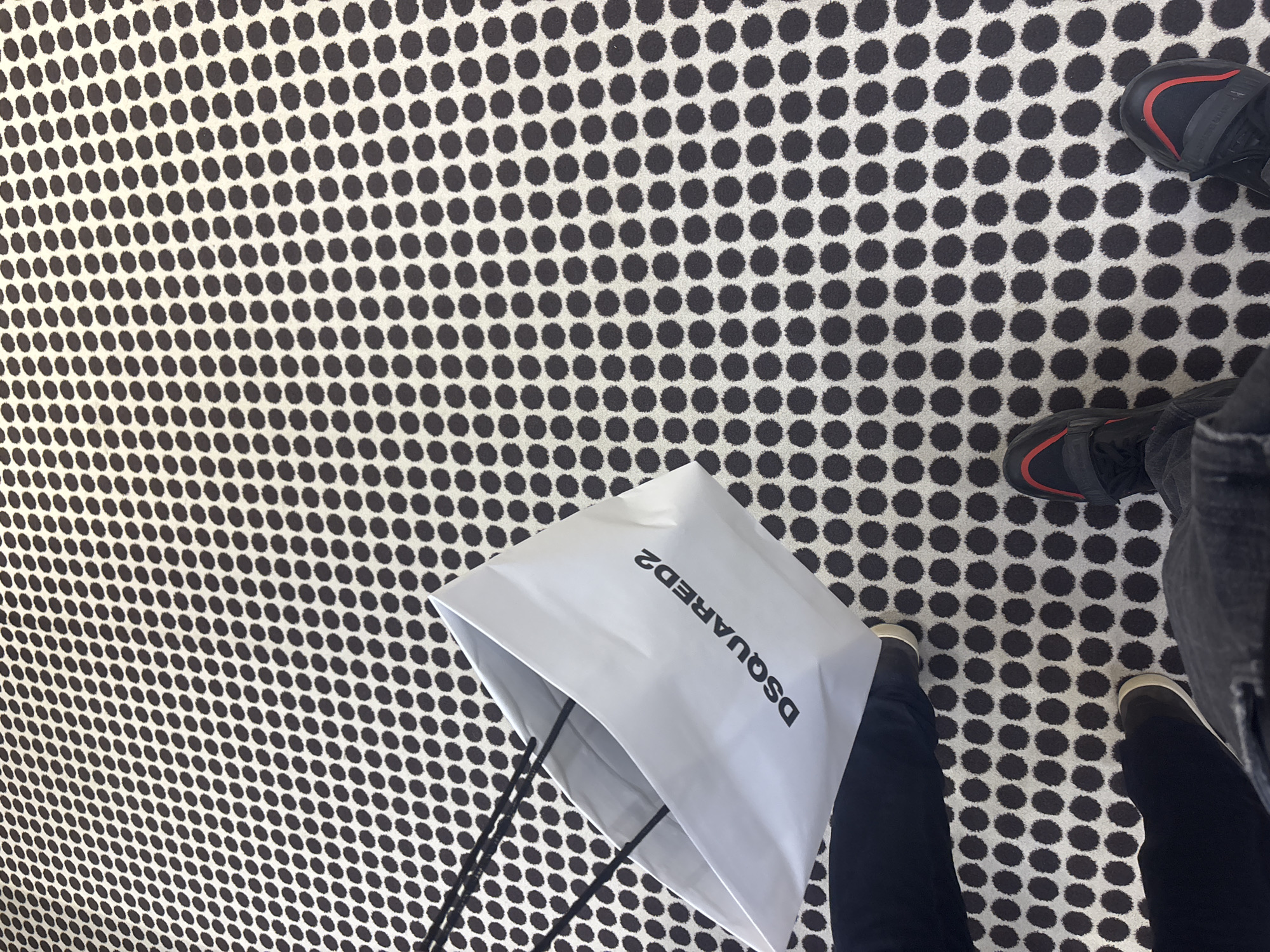 The image presents a high-angle, overhead view of a person's feet and a shopping bag on a distinctive patterned floor. The primary subjects are a white shopping bag and the person's lower legs and feet. The shopping bag features the brand name "DSQUARED2" printed prominently in black, uppercase letters. The person is wearing dark trousers or jeans. One foot, visible in the upper right, wears a black shoe with a red accent band. Another foot, visible in the lower right, wears a dark shoe with a lighter sole.
The activity depicted is shopping, suggested by the presence of the branded shopping bag in what appears to be a retail environment. The setting is indoors, as indicated by the uniform flooring and lack of outdoor elements.
A notable detail is the floor's pattern, which consists of numerous evenly spaced black polka dots against a light, possibly off-white, background. This repetitive geometric design covers the entire visible floor area. No specific time of day or weather conditions are discernible. The provided location context is Mendrisio, Switzerland.