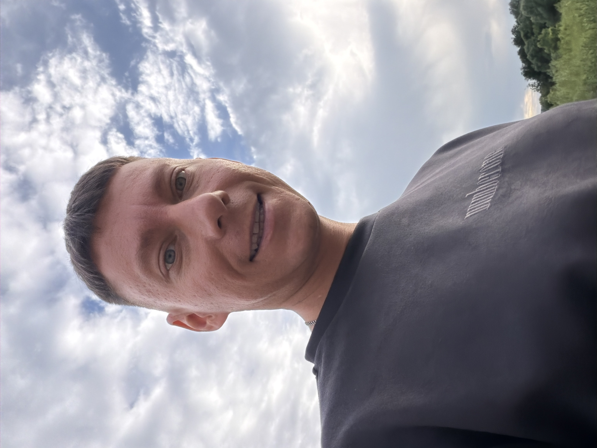 This is a selfie of a man taken from a low angle, looking up at the sky. The man has short brown hair and is smiling. He is wearing a dark t-shirt with a white logo on the sleeve. The background is a cloudy sky, with patches of blue visible. To the right, there is a blurry green landscape, possibly trees and grass. The lighting suggests it is daytime, likely overcast. The image appears to have been taken outdoors. The logo on the t-shirt contains white, diagonal lines forming a pattern. There is no other visible text. The location is identified as Moscow, Russia.