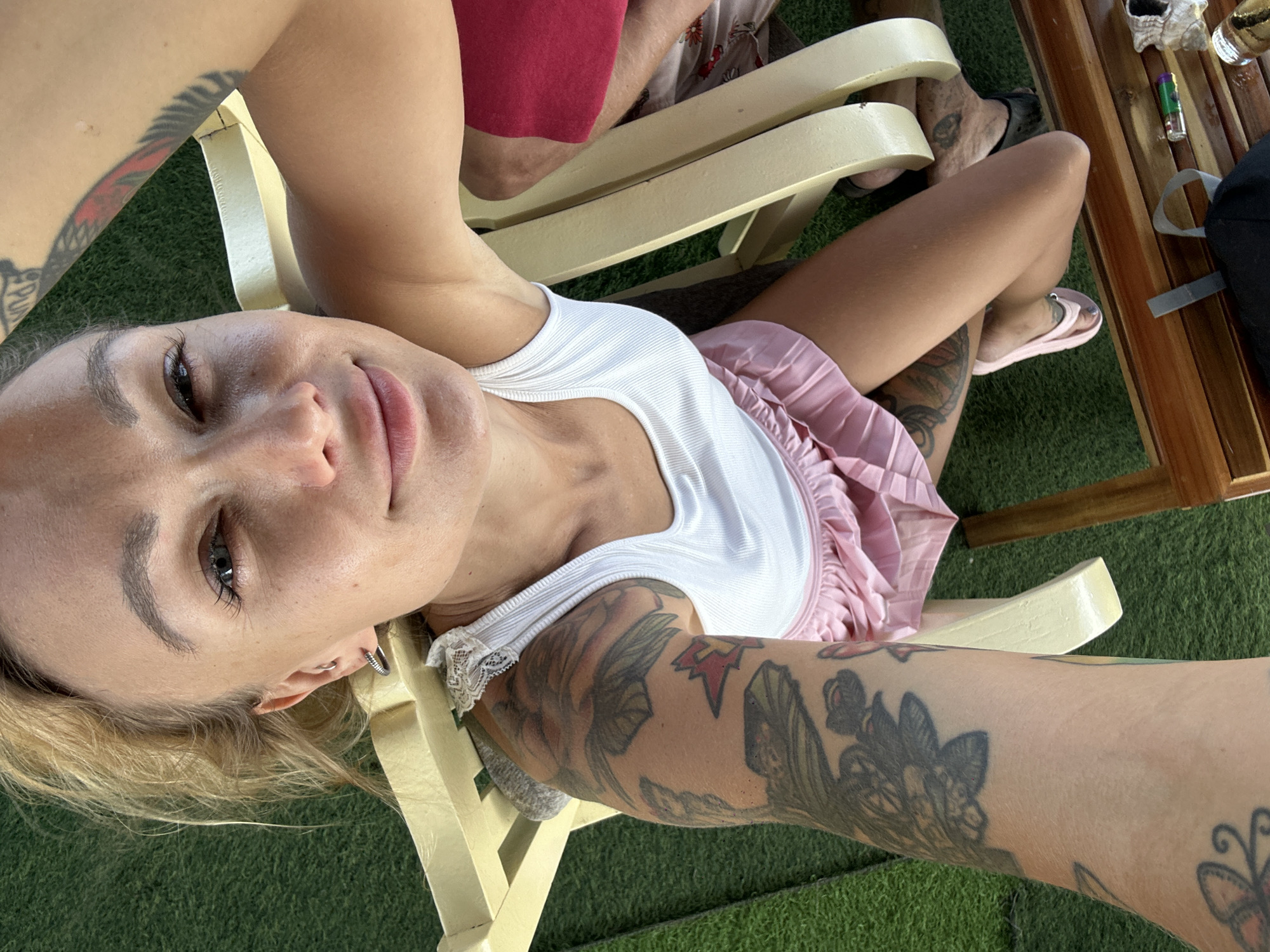 A high-angle selfie photograph captures a woman sitting outdoors in Phu Thuy Ward, Vietnam. The woman, with blonde or light brown hair pulled back, looks directly into the camera with a subtle smile. She has defined eyebrows, visible eyelashes, and small silver hoop earrings in both ears. She wears a white ribbed tank top with lace trim on the straps and a pink pleated mini-skirt. Her left arm, prominently featured in the foreground, is heavily tattooed with various designs including floral and avian motifs. Additional tattoos are visible on her right upper arm and left thigh. On her left foot, she wears a light pink ballet-style flat shoe. She is seated on a light yellow wooden slatted chair.
In the background, the ground is covered with artificial green turf. To the right of the primary subject, the legs of at least one other individual are partially visible, clad in red shorts and dark footwear, suggesting another person is seated nearby on a similar yellow chair. In the lower foreground, a dark brown wooden table or bench is visible, holding a black bag strap, a small green and purple lighter, a crushed white paper item, and a silver-colored metal object. The scene is brightly lit by natural daylight.