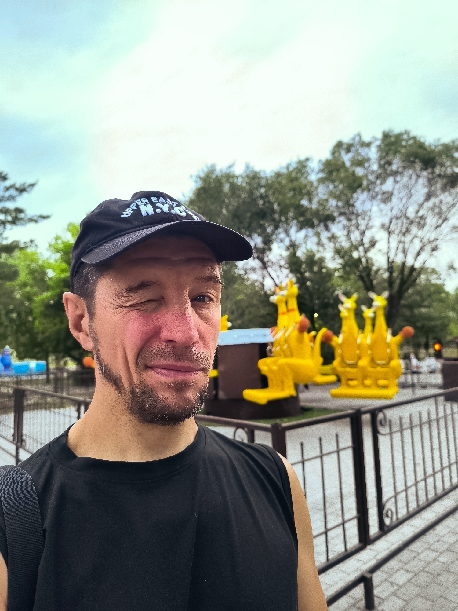A man wearing a black baseball cap with "UPPER EAST N.Y.C." visible on it, and a black sleeveless shirt, is taking a selfie outdoors. He is winking, and his right cheek is flushed red. He has short brown hair and a goatee. Behind him, there is an amusement park ride featuring multiple bright yellow kangaroo figures. A metal fence with a black railing encloses the ride area. Trees and a cloudy sky are visible in the background. The location appears to be an outdoor amusement park in Qarağandı city administration, Kazakhstan.