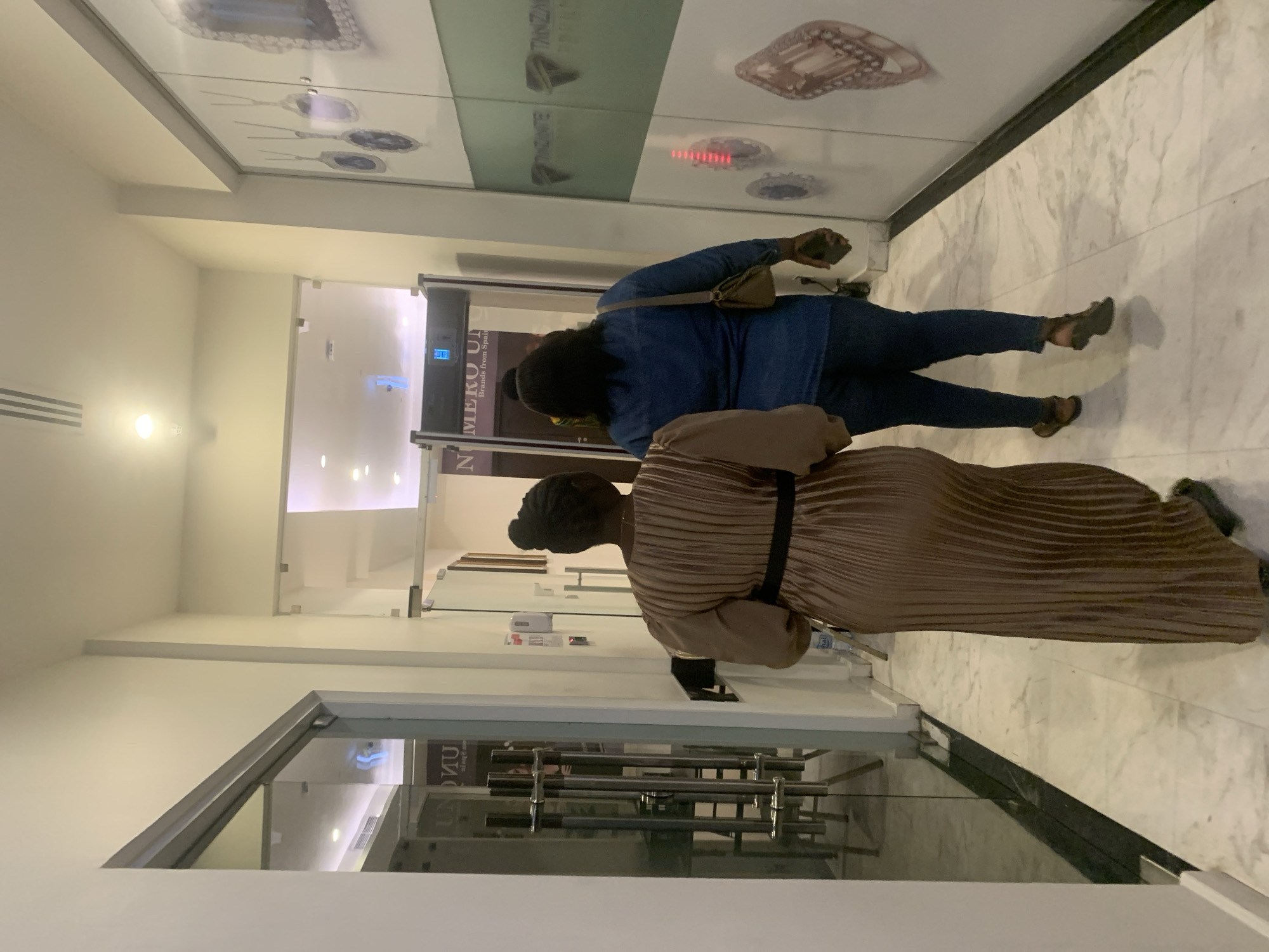 Two women are standing in front of a store in Dar es-Salaam, Tanzania. One woman is wearing a blue shirt and jeans, and the other is wearing a brown dress with a black belt. The store has large windows with images of gemstones and the word "TANZANITE" on them. The storefront has the words "NUMERO UNO Brands from Spain" visible in the glass doors. The floor is marble. The area appears to be an upscale shopping mall.