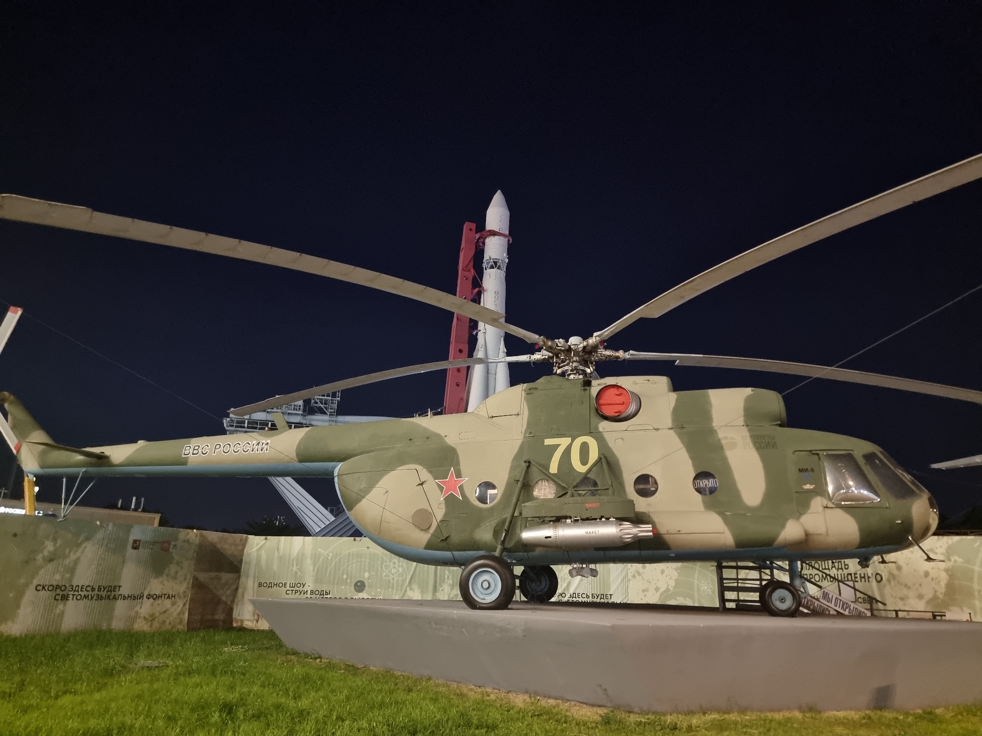 This image captures a large, camouflaged helicopter on display at night, with a rocket in the background. The helicopter, which appears to be a Mil Mi-8, is painted in a green and tan camouflage pattern. It has large rotor blades, a red star emblem on its side, and the number "70" painted in yellow. A cylindrical object, possibly a missile or fuel tank, is mounted underneath the fuselage.
Behind the helicopter, a white rocket with red accents stands vertically. The rocket has Cyrillic lettering on its side. The scene is set against a dark night sky.
The lower part of the image shows a grassy area and a concrete display platform. There are informational panels with text in Russian, some of which are partially visible. One panel has text that reads "СКОРО ЗДЕСЬ БУДЕТ СВЕТОМУЗЫКАЛЬНЫЙ ФОНТАН" (Soon there will be a light and music fountain here) and "ВОДНОЕ ШОУ - СТРУИ ВОДЫ" (Water show - water jets). Another panel has text that is difficult to read but appears to mention an industrial area.
Based on the presence of the rocket and the display of Soviet-era military hardware, this scene is likely located at the All-Russian Exhibition Center (VDNKh) in Moscow, Russia. VDNKh is a permanent national exhibition in Moscow featuring many pavilions and outdoor displays, including a space exploration exhibit that often features rockets and aircraft.