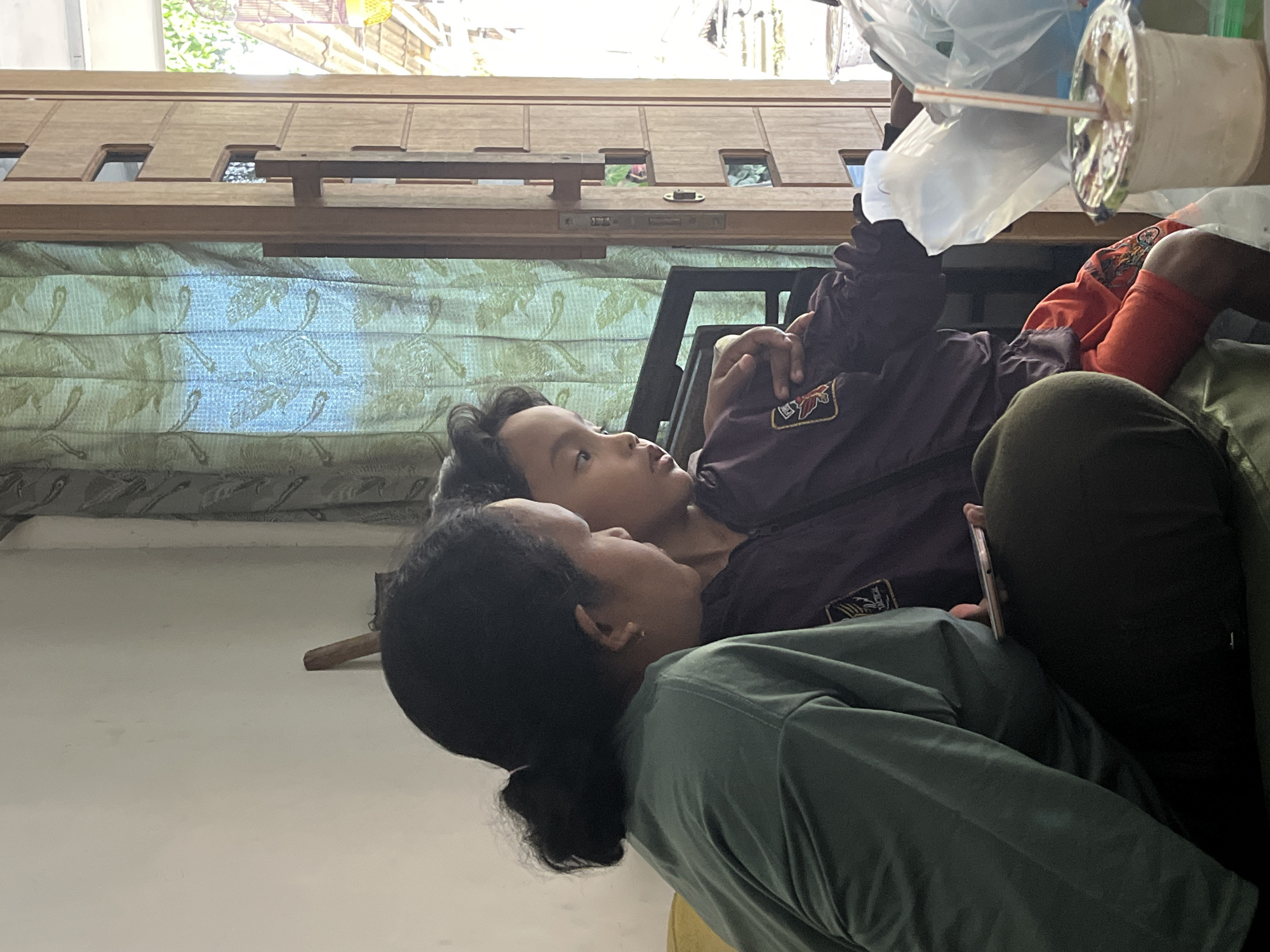 In an indoor setting, likely a home, two individuals are the central focus. On the left, a person with dark hair pulled back in a bun, wearing a dark green long-sleeved shirt, is seated. Their arm is bent, and they appear to be holding a smartphone. To their right and slightly in front, a child with dark, curly hair is also seated. The child is wearing a dark purple jacket with an emblem on the left chest, and bright orange shorts. The child is looking towards something off-camera to the right. The two individuals are positioned closely, suggesting an interaction, possibly shared viewing of the smartphone. The background includes a curtain with a patterned design, a wooden door, and glimpses of an outdoor area with sunlight. In the foreground, to the right, a plastic cup with a straw and a lid is visible, along with plastic bags. The overall impression is a candid moment captured in a domestic environment. The location is identified as Pasrukkrajan Satu, Indonesia.