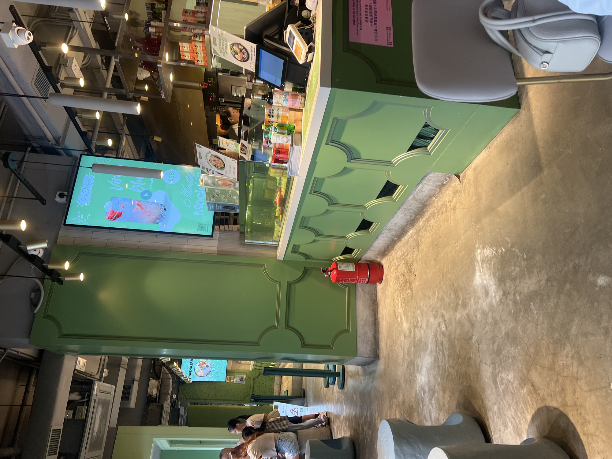 The image depicts the interior of a modern, brightly lit establishment that appears to be a cafe or restaurant in Hong Kong, China. The color palette is dominated by vibrant green, seen in the decorative paneling on a pillar and the counter area, contrasted with the neutral tones of the concrete floor and light-colored seating.
In the background, a large digital screen displays a vibrant image of a drink with the text "SourDough" and "$68", suggesting promotional content for the establishment. Behind the counter, a person is visible, seemingly engaged in work. The counter itself is lined with various bottles of colorful drinks and other merchandise. A red fire extinguisher stands prominently near the green counter, a standard safety feature in public spaces.
In the foreground and mid-ground, there are light blue, tree-stump-like stools and a grey chair with a handbag placed on it. The flooring is a polished concrete with visible cracks and some white dust or residue. The lighting fixtures, including spotlights and hanging lamps, contribute to the bright and contemporary atmosphere. The overall impression is of a clean, organized, and bustling commercial space within an urban environment.