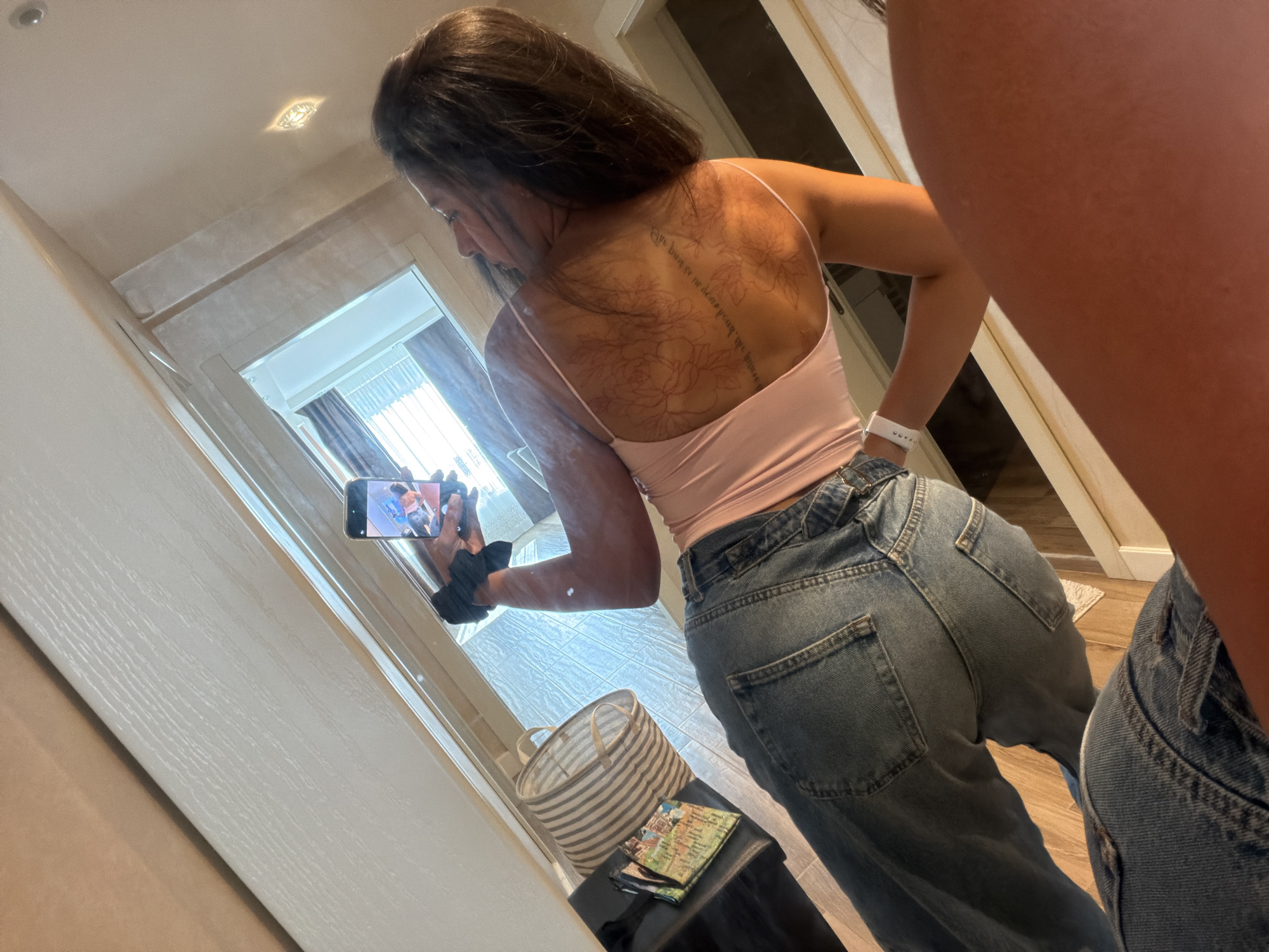 A young woman with dark hair is taking a mirror selfie. She is wearing a pink spaghetti strap crop top and blue denim jeans. Her back is visible, revealing a large tattoo of flowers and script across her upper back. She is holding a smartphone in her right hand, and a black hair scrunchie is attached to her left wrist. To her left, a striped laundry basket and a stack of postcards are visible on a dark surface. The scene appears to be indoors, possibly a hotel room, with reflective surfaces and bright lighting. The background shows a window with blinds and a glimpse of an outdoor view. This image is not located in Gabrovo, Bulgaria.