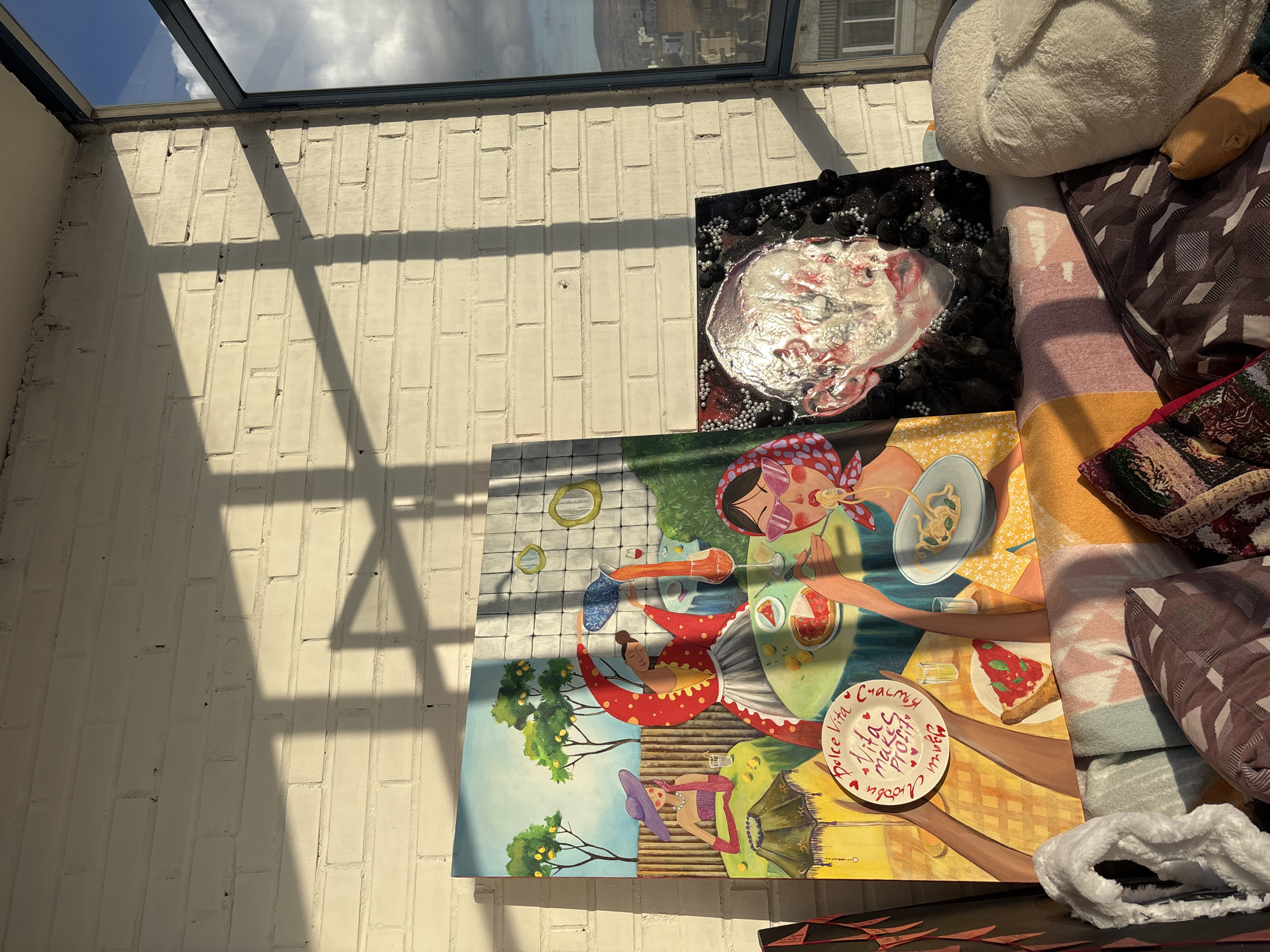 The scene depicts a sunlit interior space in Krasnoyarsk, Russia. A section of a white brick wall is visible, intersected by shadows cast from a window frame or skylight above. Two paintings lean against the wall or are displayed near it. One painting is a mixed media piece with a textured, possibly metallic, representation of a head or face with black and silver embellishments on a black background. The second painting features an illustration depicting leisurely scenes including women in various outdoor settings, one involving a picnic or meal with the words "Dolce Vita makes project Vita счастье" on a plate. Adjacent to the paintings are a pile of soft goods: a white cushion, a striped blanket, and various patterned pillows. Part of a window is visible at the top of the frame, showing a cityscape or landscape. The environment appears to be a bright, possibly indoor, studio or living space.
