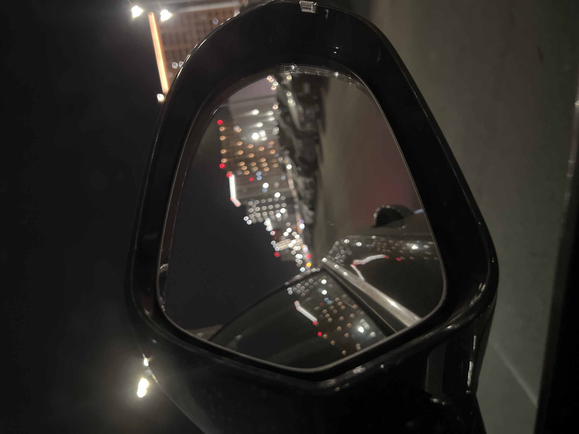 A black car side mirror shows a night scene reflecting city lights. The reflection contains blurred lights from tall buildings and streetlights, indicative of a dense urban environment. To the left of the mirror, the background is dark, potentially indicating a dark sky or a poorly lit area. The location is presumed to be Tyumen, Russia based on the request.