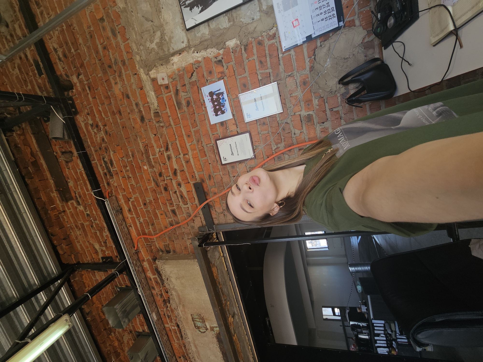 The image appears to be a selfie taken by a woman indoors, featuring a background with a brick wall adorned with framed documents and a calendar. A desk with various items, including a telephone and a purse, is also visible. The woman is wearing a green top with what looks like a book or magazine on the front. The location seems to be an industrial-style space with visible metal structures. Based on the OCR text, the location is likely in Chaikovsky, Russia.