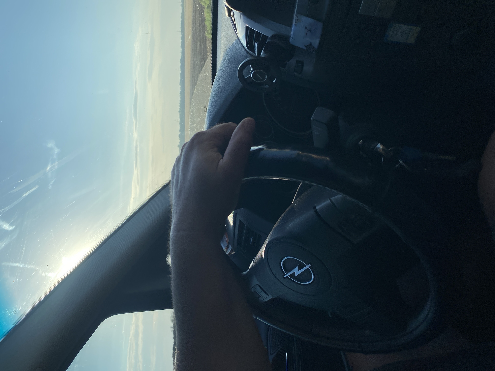 An over-the-shoulder shot captures a person's left hand resting on the steering wheel of a car, as seen from the driver's perspective. The steering wheel has the Opel logo embossed on it. Beyond the windshield, a rural landscape unfolds under a bright, sunny sky with scattered clouds. The horizon features fields and what appear to be utility poles. The scene is set on a road outside of Maiske, Ukraine. The interior of the car shows part of the dashboard and a small, circular accessory attached to it.