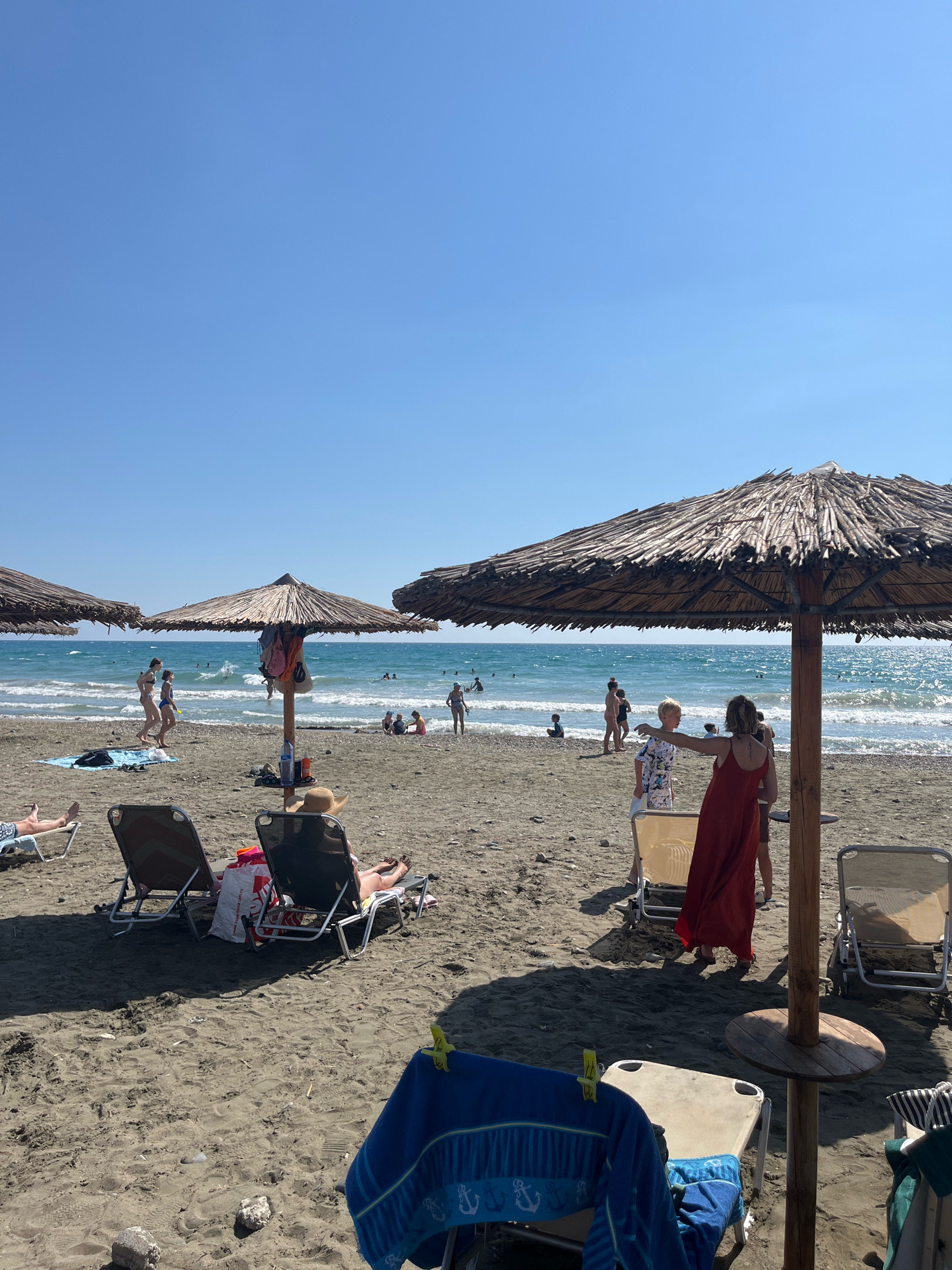The image depicts a sunny day at Episkopi Beach in Episkopi, Limassol, Cyprus. The beach is sandy, with several thatched umbrellas providing shade for beachgoers. In the foreground, there are lounge chairs with towels draped over them. In the middle ground, people are seen walking on the beach and in the shallow water. The sea is calm with gentle waves. In the background, the clear blue sky extends to the horizon. The overall scene conveys a relaxed atmosphere of a typical summer day at the beach.