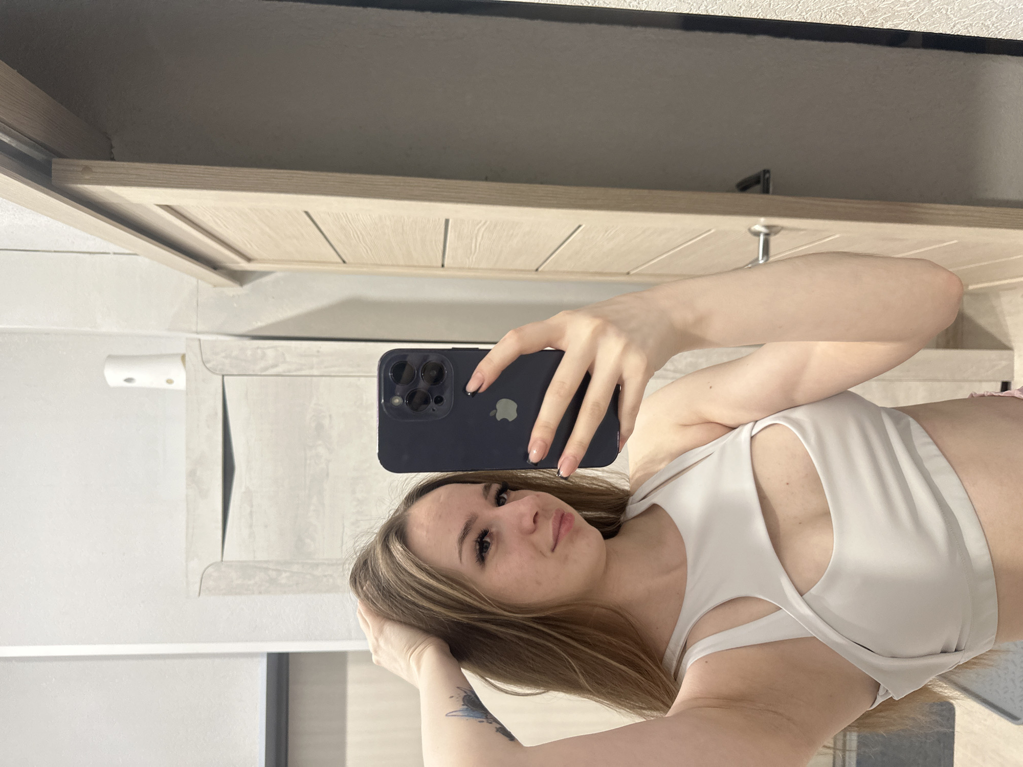 Here is a detailed description of the image:
A young woman with blonde hair is taking a selfie in a room. She's holding a dark-colored smartphone in her right hand, partially obscuring her face. She's wearing a white sports bra that reveals part of her chest. A tattoo is visible on her left arm.
The background consists of a light-colored wooden cabinet and what appears to be a white wall with a built-in shelf. The setting is indoors and seems to be in a domestic environment.