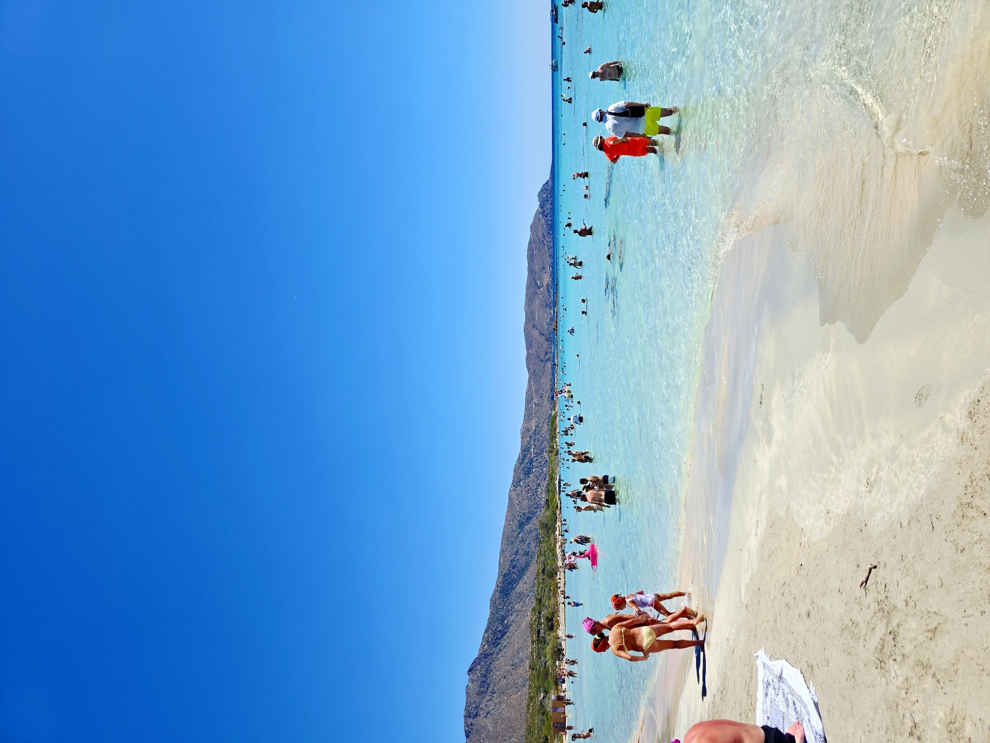 This media file shows a lively beach scene under a clear blue sky, suggesting a sunny day. The image is oriented vertically. The primary subjects are a multitude of people enjoying a tropical beach. The setting is a wide, sandy beach with crystal-clear, turquoise water lapping at the shore. In the background, a rugged, mountainous landscape rises behind the beach.
Numerous people are scattered across the scene, engaged in various beach activities. Some are wading in the shallow water, while others are swimming or standing in the sea. Several individuals are visible on the sand, some appearing to be relaxing or walking. Close to the foreground, three people, two women and a child, are interacting at the water's edge. One woman wears a yellow bikini, and another wears a white swimsuit. A child in the middle is also in swimwear. Further back, more people are seen in the water, including a man in a red life vest, and a group of people near the shore, with some appearing to be tourists.
The water is remarkably transparent, allowing for a clear view of the sandy bottom in the shallower areas. Gentle waves are breaking on the shore, creating a delicate white foam. The sand is light-colored and appears fine-grained. The overall atmosphere conveyed is one of leisure and summer fun, with people actively enjoying the warm weather and the sea. There is no visible text in the image.