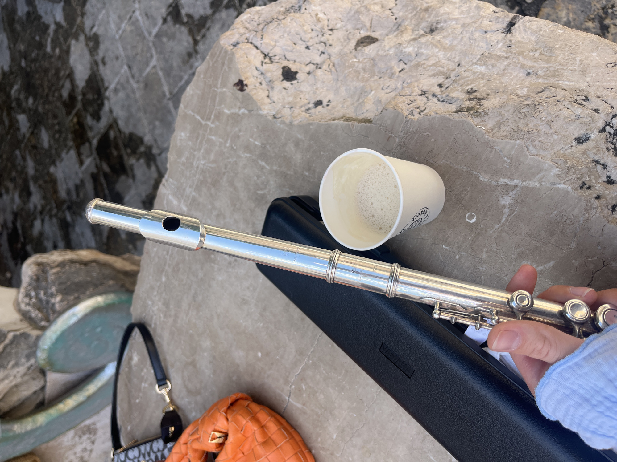 A silver flute lies on a black flute case, next to a cup of coffee, atop a light-colored stone surface. Adjacent to the flute case, there is a small black and white purse with a gold strap and an orange, textured handbag. A person's hand, clad in a light blue sweater, holds the end of the flute. The background shows textured stones and a turquoise-painted rock formation. The image was likely taken in Budva, Montenegro.