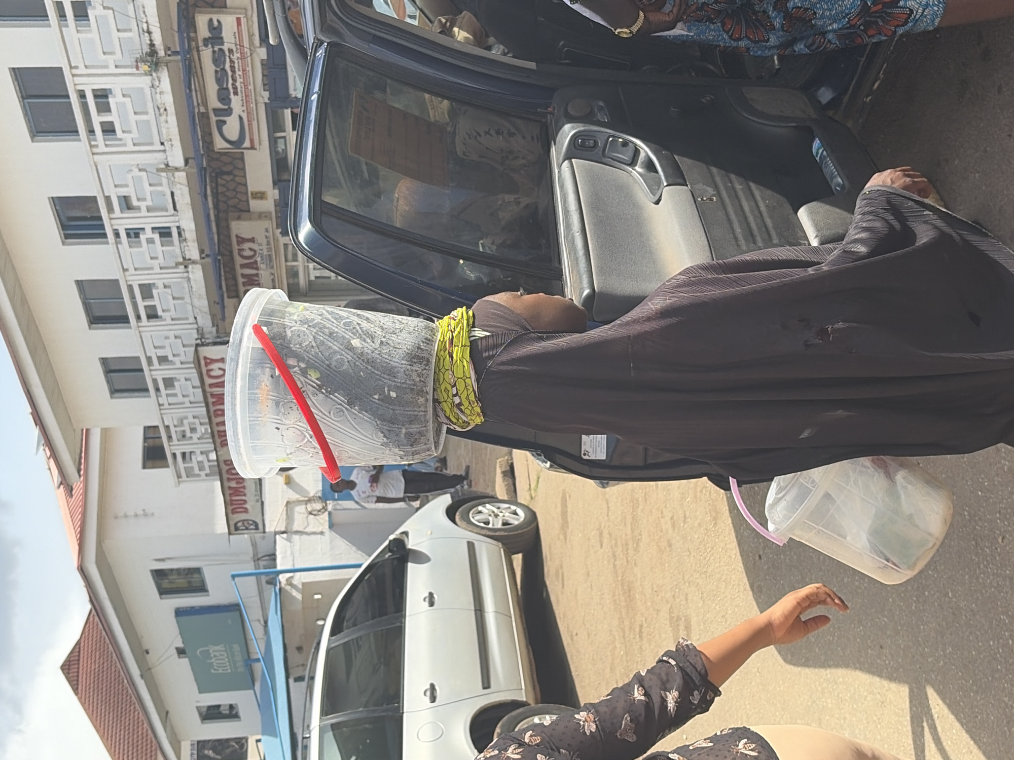 The image shows a street scene in Jos, Nigeria, with a person wearing a black abaya and a yellow and green headscarf. They have a large, clear plastic bucket on their head, filled with what appears to be dark material, possibly food items or produce. A red handle sticks out from the bucket. The person is leaning into the open door of a dark blue vehicle, suggesting they might be placing or retrieving the bucket. Another person, partially visible from the shoulders down, is wearing a patterned top with blue and orange motifs and a gold watch. Their hand is reaching towards the car door.
In the background, a white van is parked on the street. Behind the van, a multi-story building is visible, with the signs "Classic" and "PHARMACY" displayed prominently. Another sign reads "DUMJOZ PHARMACY." The architecture of the building suggests an urban setting. The lighting indicates it is daytime, with shadows cast on the ground, implying a sunny day. The overall scene depicts an everyday activity of transporting goods, likely in a marketplace or residential area.