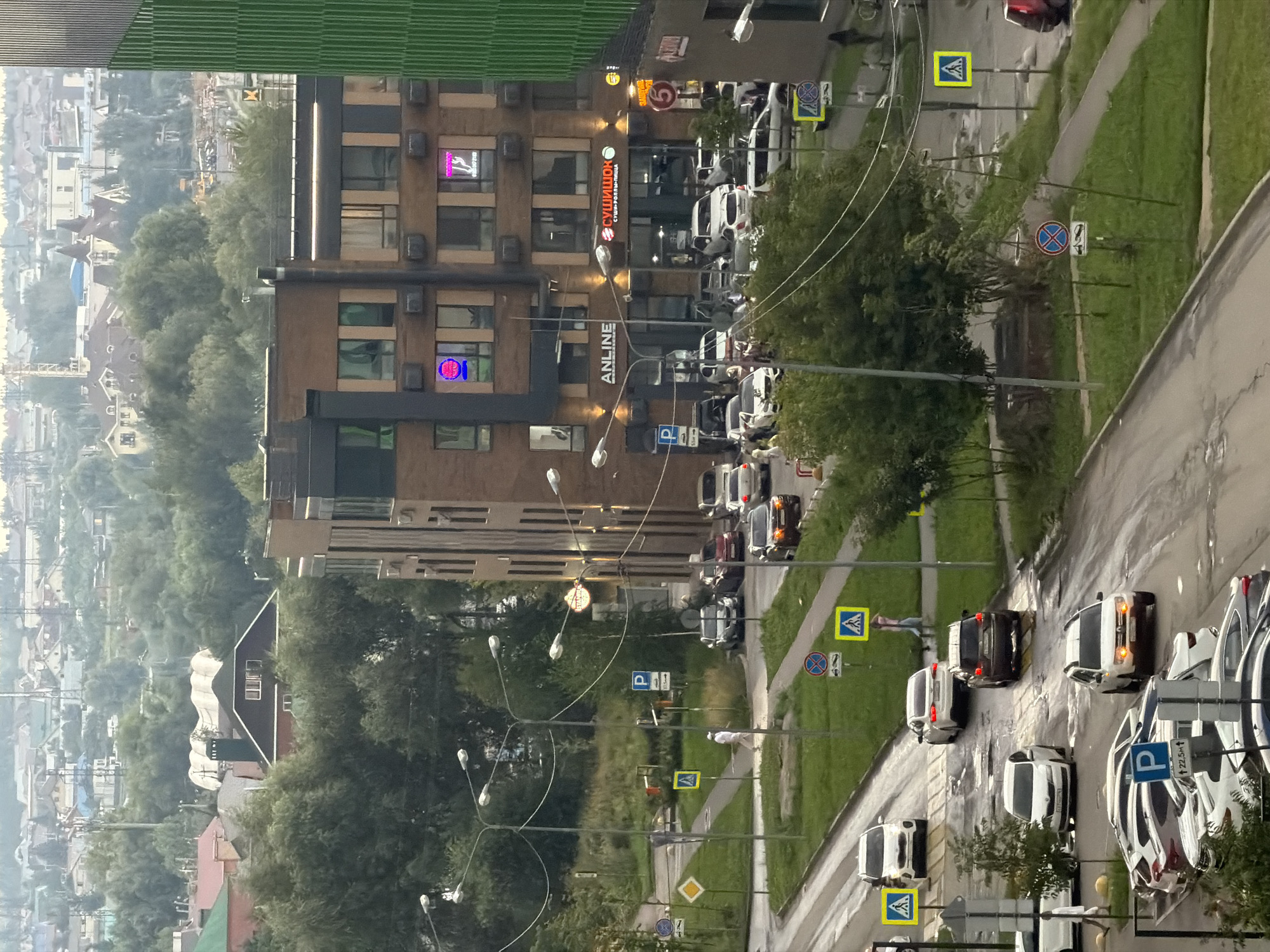 The image shows a street scene in Kazan, Russia, with a multi-story brick building on the right side of the frame. The building has storefronts on the ground floor, including one with a neon sign that reads "ANLINE" and another with a red sign that says "СУШИШОК" (Sushi shop). Several cars are parked or driving on the wet street, which appears to have puddles from recent rain. Pedestrian crossings with yellow warning signs are visible. People are walking on the sidewalks and crossing the street. The background shows a densely wooded area and a distant cityscape with residential buildings. A tall green structure is partially visible on the far right edge of the image.
