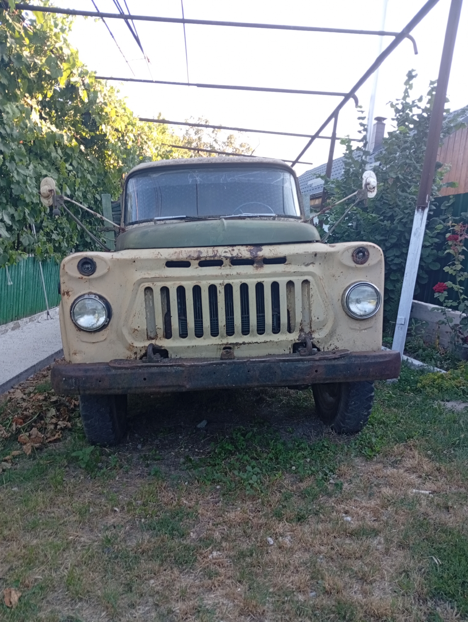 An aging, light-colored truck with a green upper body is parked on a grassy area in Pănășești, Moldova. The truck is the primary subject, positioned centrally and facing forward. Its front end exhibits signs of wear, with rust visible on the bumper and surrounding areas. The headlights are round, and the grille is vertical. A metal structure, possibly a canopy or frame, extends over the truck from the top of the image. In the background, there is dense foliage, a wooden fence, and a building with a dark roof. The ground is covered in a mix of dry and green grass with scattered fallen leaves. The overall environment suggests an outdoor, possibly rural or semi-urban setting.