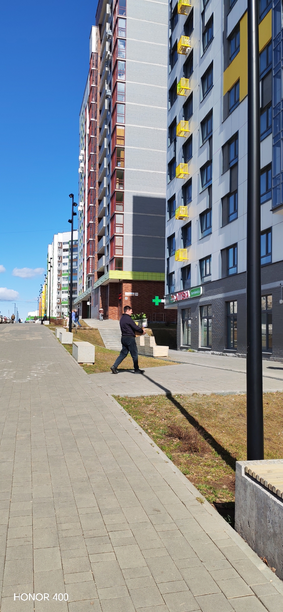 A man carrying a basket walks along a paved sidewalk on a sunny day in Izhevsk, Russia. The man is wearing a dark jacket and dark jeans. He is walking to the left. The sidewalk is made of square grey pavers. To the right of the sidewalk is a patch of dry grass. A black light pole stands to the right of the sidewalk, casting a shadow. Further to the right is a concrete bench with a wooden seat. In the background are several tall apartment buildings. The building closest to the man has a pharmacy ("APTEKA") on the ground floor, marked with a green cross. The address "10" is also visible on the building. The buildings are painted in a variety of colors, including grey, red, yellow, and green. The sky is blue with a few scattered clouds. There are a couple of people walking in the distance. Cars are parked far in the distance. A "HONOR 400" watermark is at the bottom left of the image.