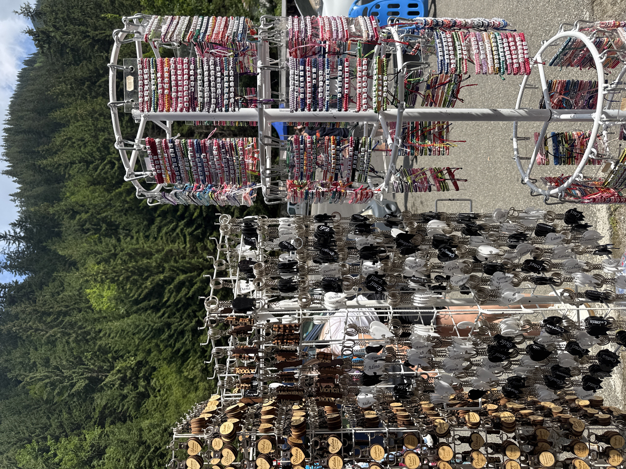 This is a medium-shot, outdoor, eye-level photograph of a street market stall in Cârțișoara, Romania. The stall displays a large selection of personalized keychains and bracelets. The keychains, predominantly wooden and metal, are arranged in vertical rows on the left side of the frame. They are round and heart-shaped, some with engraved names and designs. The bracelets, displayed on a rotating rack on the right, are made of colorful threads and beads, with individual letter beads spelling out various names. A person in a white shirt is partially visible behind the keychains, and another person wearing dark clothing is visible in the background, near a blue cart. The market is set against a backdrop of dense, green pine forest and a bright, partly cloudy sky. The ground appears to be asphalt.