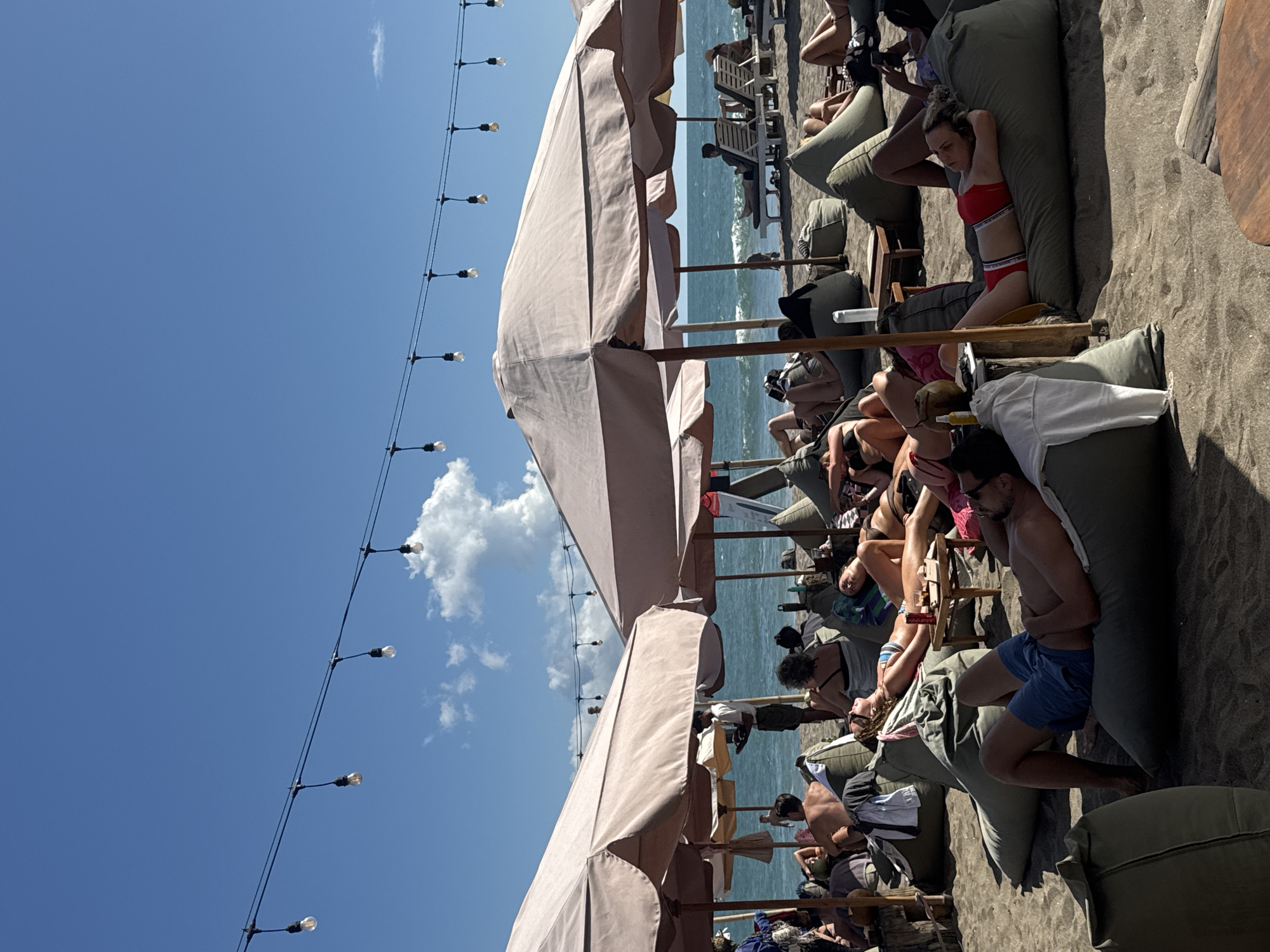 This image shows a beach scene in North Kuta, Indonesia. Several people are relaxing on bean bag chairs and lounge chairs under large umbrellas on a sandy beach. Some are sunbathing, while others are conversing. The ocean is visible in the background, and a string of lights hangs overhead. The sky is blue with a few clouds.