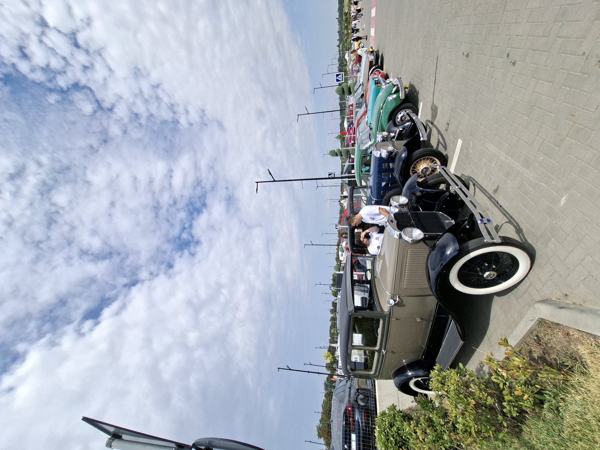 The image depicts an outdoor event, likely a car show, in the city of Stăuceni, Moldova. A row of vintage cars is parked on a paved surface, with a blue sky and scattered clouds overhead. The foreground is dominated by the front portion of a tan and black vintage car with white-walled tires. Behind it, several other classic automobiles are visible, including a dark blue car, a green car, and a red car. People are casually observing the vehicles. In the background, there are more cars, people, streetlights, and some greenery. The overall scene suggests a public gathering focused on classic automobiles.