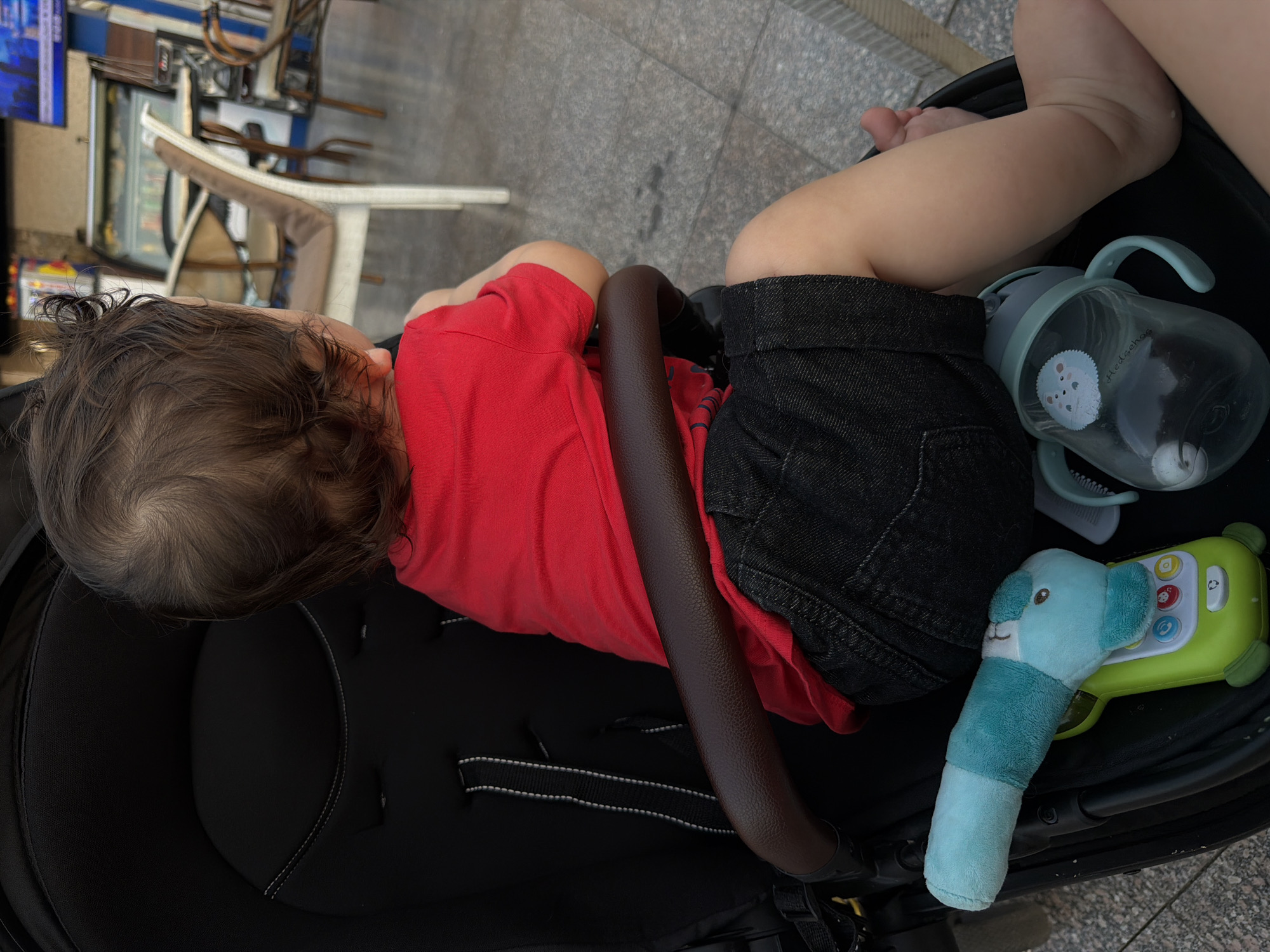 The image depicts a young child, viewed from the back, seated in a stroller. The child is wearing a red t-shirt and dark shorts. In the stroller, a blue stuffed toy resembling an animal's head and a sippy cup with a hedgehog design are visible. The stroller is positioned outdoors, with a cobblestone-like ground surface and the blurred background suggesting an urban setting with tables and chairs, possibly a cafe or restaurant. The lighting indicates daytime. The scene could be located in Sharm El Sheikh, Egypt, given the typical ambiance of outdoor seating areas in tourist locations there.
