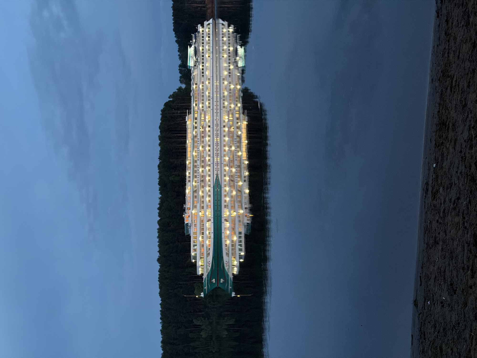This image shows a reflection of a large, illuminated cruise ship in calm water. The ship is white with multiple stories and numerous windows with lights on, suggesting it is nighttime. The ship's bow is green and white, and the name "ВЛАДИМИР КЯККОЙСКИЙ" is visible on the side. A dark line of trees runs along the edge of the water, mirroring the reflection on the other side. The sky is a dark blue, indicating dusk or night. A strip of land is visible in the lower right corner. The location is Chaikovsky, Russia.