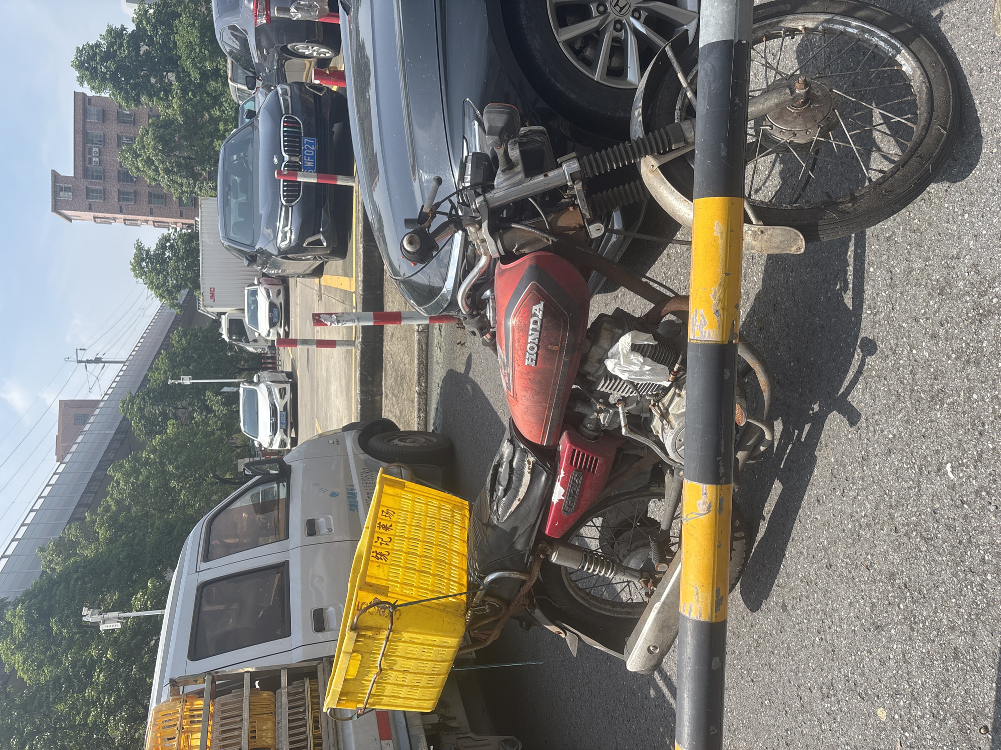 In Nanhai District, China, a weathered red and silver Honda motorcycle with a yellow plastic basket attached to its rear rack is parked on an asphalt surface. The basket appears to be used for transport, with some handwritten Chinese characters visible on its side. Behind the motorcycle, a dark-colored sedan is parked, and further in the background, a white van loaded with more yellow baskets is visible. The scene suggests a commercial or delivery context. A metal barrier with yellow and black stripes runs across the foreground, partially obscuring the motorcycle's front wheel. In the background, there are trees, elevated structures, and buildings. The sky is clear and bright, indicating daytime.