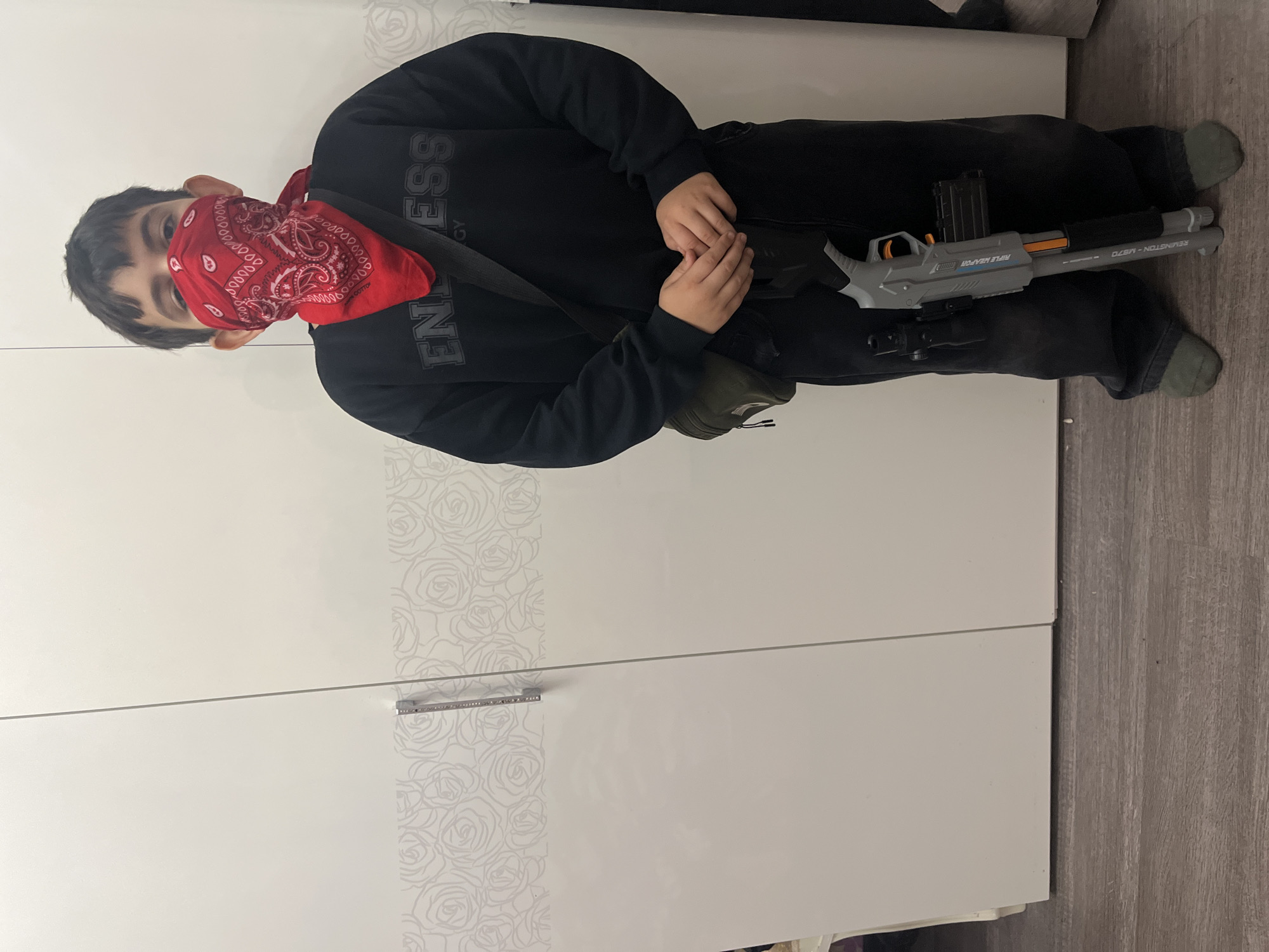 A young boy stands in front of a white wardrobe with a floral pattern. He is wearing a black sweatshirt with the word "ENDLESS" in large, dark lettering. A red bandana covers his lower face, and he has a dark green crossbody bag slung over his left shoulder. In his right hand, he holds a gray toy rifle with orange accents. The floor is a light-colored wood laminate. There is no indication of the city of Kostroma, Russia in this image.