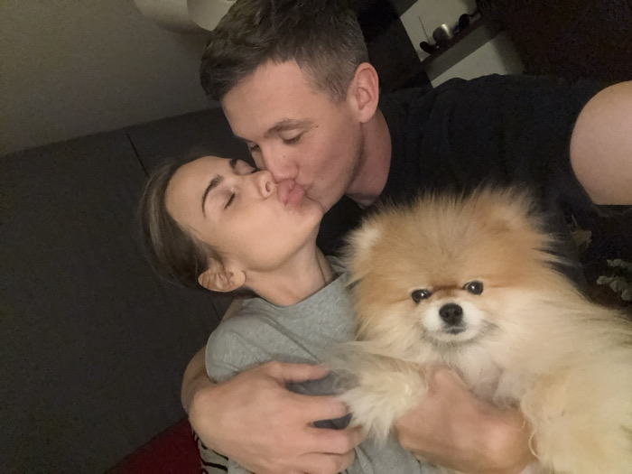 The image depicts two individuals and a dog in an indoor setting. A male subject, with dark hair and wearing a black short-sleeved shirt, is positioned on the right. He is shown kissing the left cheek of a female subject, whose eyes are closed and lips are pursed. The female subject, with light brown hair and wearing a grey t-shirt, is positioned on the left. The male's left arm is wrapped around the female. In the foreground, between and below the two individuals, a small, fluffy, light brown Pomeranian dog is held, looking directly at the camera. Both individuals appear to be interacting with the dog. The background is dark and out of focus, indicating an interior space with dim lighting; no specific details are discernible beyond vague shapes suggesting furniture or walls. The specific location within Warsaw, Poland, cannot be determined from the visual evidence presented in the image.